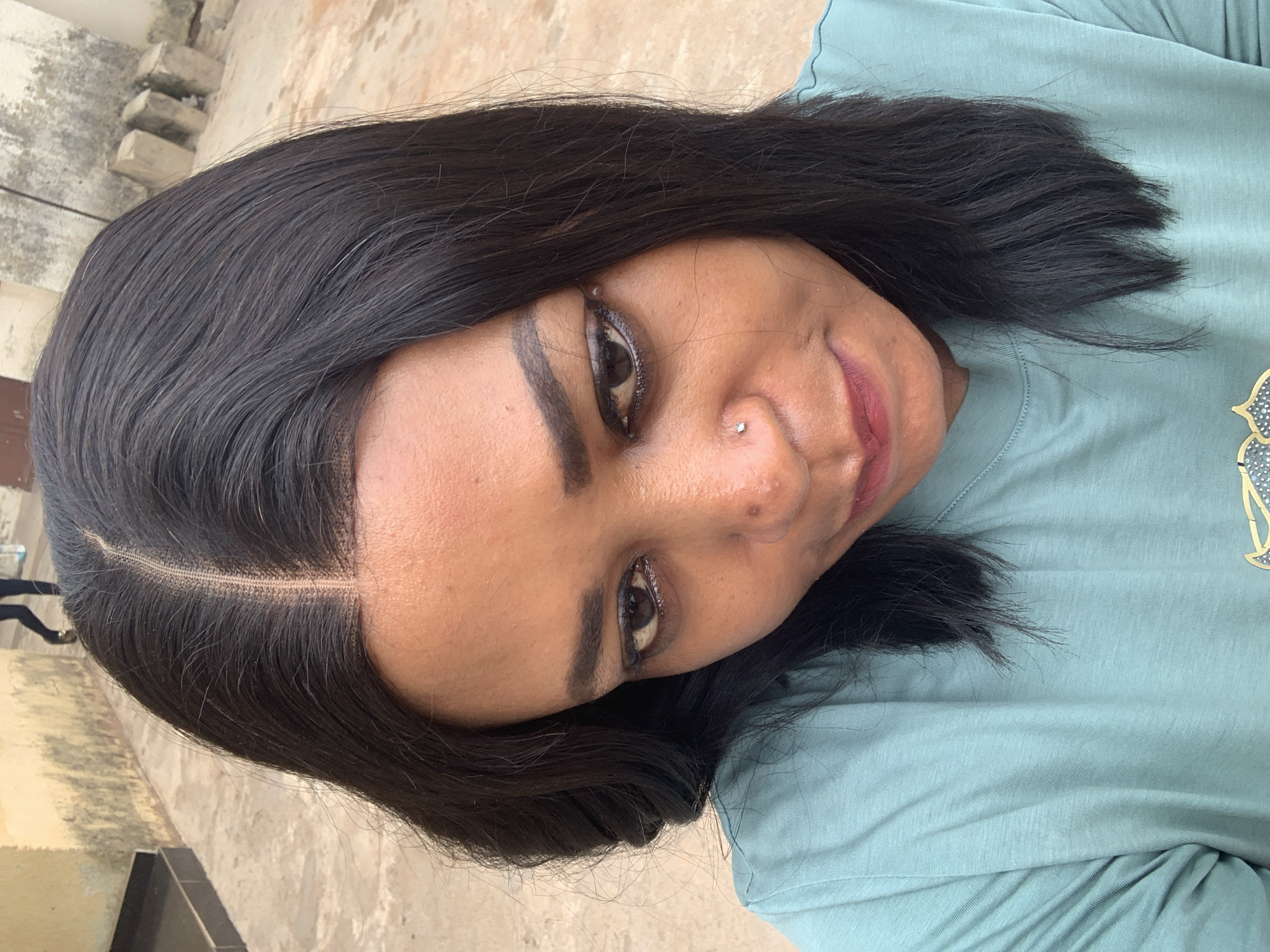 A medium-close-up, rotated headshot shows a dark-haired woman with medium skin, wearing a light blue t-shirt. The woman has dark eyebrows and eyeliner. A silver nose piercing is visible on her left nostril. The background appears to be an outdoor, sunlit area. The location is Sabon Tasha, Nigeria. The environment is a concrete surface with possible bricks.