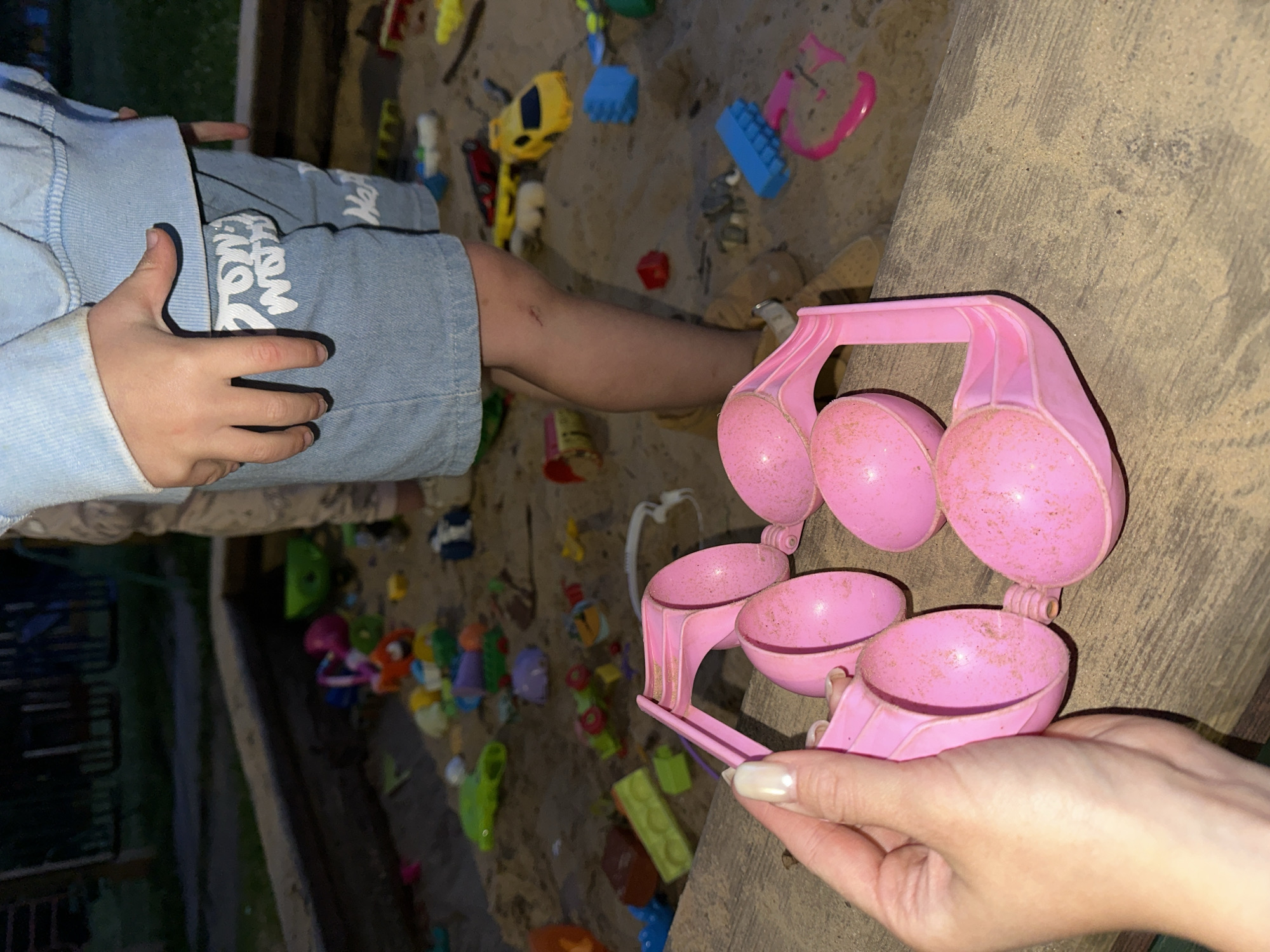 A child's hand is seen in the foreground, holding a pink plastic toy snow mold with four scoops. The toy is covered in sand. The child is wearing a light blue hoodie and denim shorts with white lettering that appears to say "NEW YORK". The child's leg is visible, showing a small scrape on the knee.
The scene appears to be a sandy playground or sandbox, likely outdoors, with various other toys scattered around, including a yellow toy car, blue building blocks, and other colorful plastic toys. The ground is sandy, with some wooden elements suggesting the edge of a sandbox or raised play area. The background is blurred but shows hints of a dark fence and greenery, possibly indicating a backyard or park setting.
The lighting suggests it could be daytime, possibly late afternoon or early evening, given the shadows and overall ambient light. The presence of sand and outdoor toys points to a recreational activity for children. The focus is on the child's hand and the toy, highlighting a moment of play. No specific weather conditions are apparent, but the sand suggests a dry environment. The location context of Nizhny Novgorod, Russia, provides a geographical anchor.