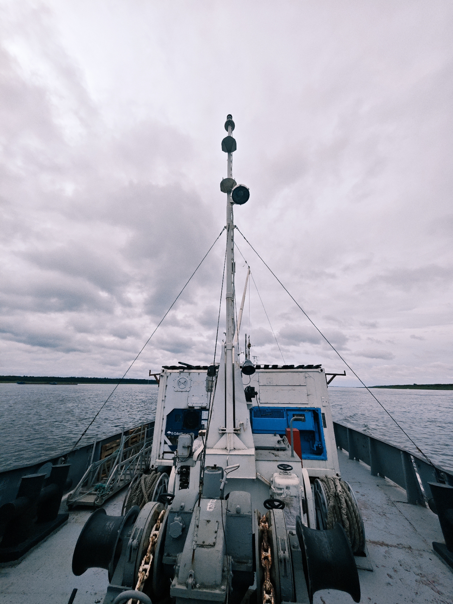 A first-person perspective shot of the deck of a ship moving through a body of water under a cloudy sky. In the foreground, large metal winches and chains are visible, with a thick rope coiled on a reel to the right. A white structure with blue and red accents rises in the midground, topped by a mast with lights. The water stretches to the horizon, where a tree line defines a distant shore under a heavy, overcast sky. The image appears to be taken on the Yenisei River near Zotino, Russia, a remote location known for its logging industry and river transport.
