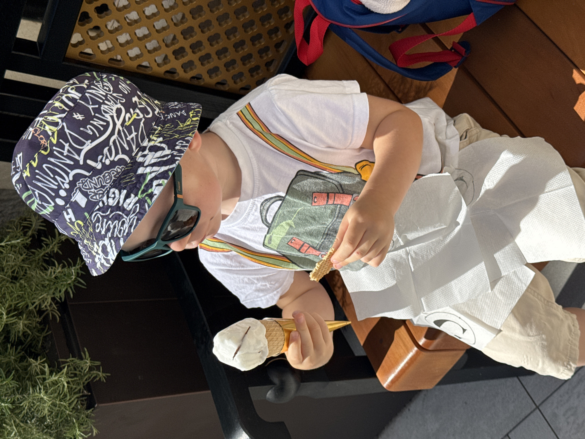 A child wearing a patterned sun hat and sunglasses sits outside on a wooden bench in an outdoor setting. The child is eating an ice cream cone, using multiple paper napkins. A blue and red backpack is on the bench next to the child. A green plant is visible in the corner of the frame, near a dark surface. The scene is sunlit. There is no visible proof of the city of Budva, Montenegro.