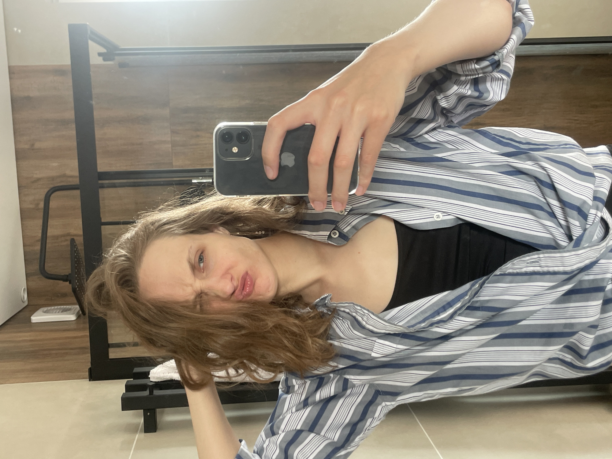 An indoor, mirror selfie captures a young woman with long, wavy brown hair, rotated 90 degrees to the left. She has a playful expression, pouting her lips and squinting one eye while looking towards the camera. She is wearing a blue, grey, and white striped button-down shirt unbuttoned over a black top. Her right hand holds a dark-colored iPhone, encased in clear plastic, prominently displaying the Apple logo and dual cameras on its back.
The background, reflected in the mirror, features wooden wall panels on the upper section and a light-colored tiled floor below. A dark metal frame structure, possibly a shelf or rack, is visible behind her. Other objects in the reflection include a white rectangular item (perhaps a scale) and a black, circular, perforated object on the floor. The lighting suggests daytime. This scene takes place in Podshivalovo, Russia.