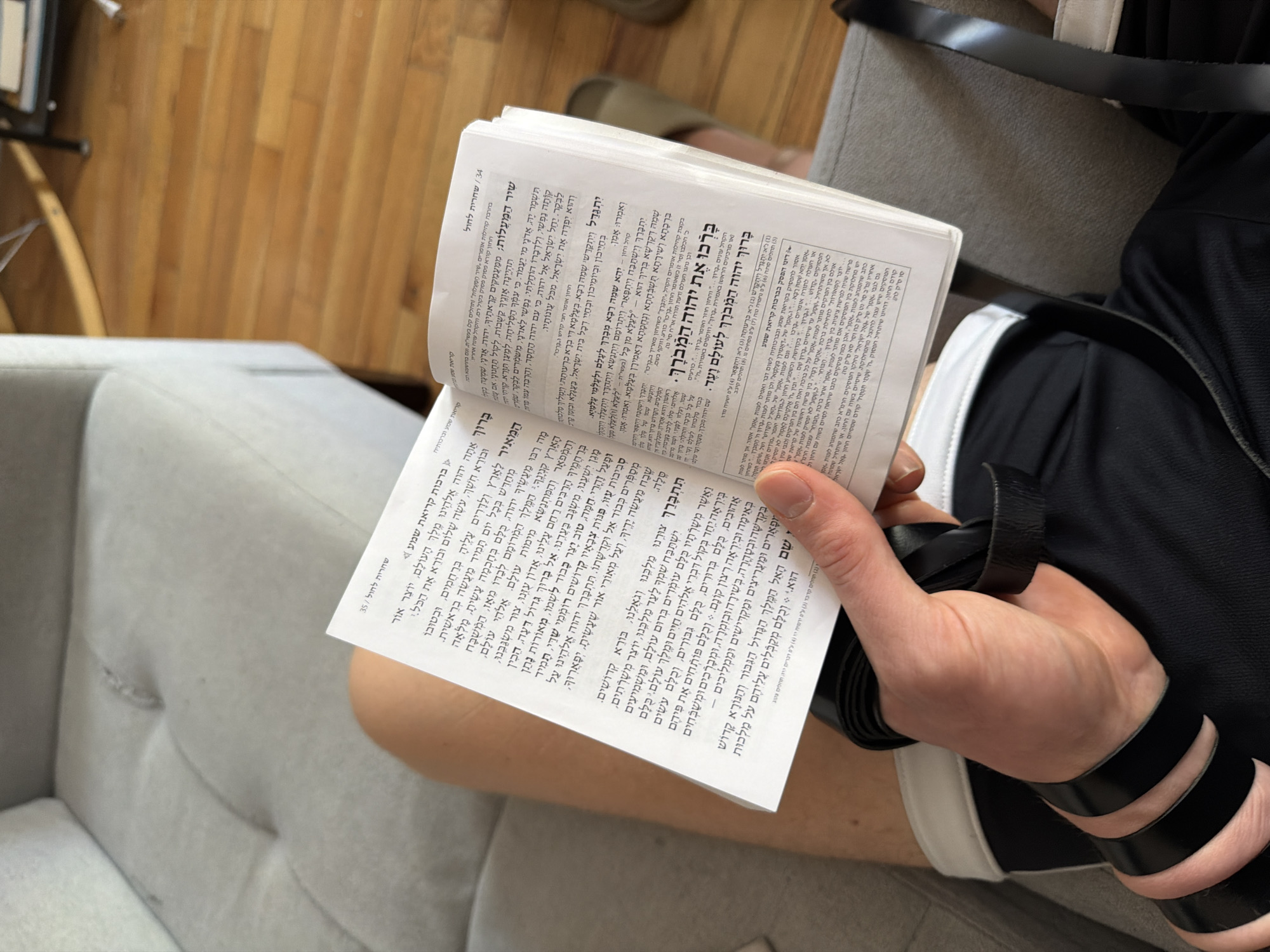 A person is shown seated indoors, likely in New York, USA, engaged in Jewish morning prayers. The individual's left arm is adorned with black leather tefillin straps, a religious observance typically worn during weekday morning services. Their right hand holds an open Siddur (Jewish prayer book), with pages 34 and 35 visible. The Hebrew text on the pages includes headings such as "שחרית לחול" (Shacharit for weekdays), "קריאת שמע" (Kriat Shema), and "ברכות קריאת שמע" (Blessings of Kriat Shema), confirming the prayer context. Specific phrases like "בָּרוּךְ אַתָּה יהוה אֱלֹהֵינוּ מֶלֶךְ הָעוֹלָם" (Blessed are You, Lord our God, King of the Universe) and "בָּרְכוּ אֶת יהוה הַמְבֹרָךְ" (Bless the Lord, the Blessed One) are discernible. A section titled "דיני הפסק בברכות קריאת שמע" (Laws regarding interruptions during the blessings of Kriat Shema) provides halachic guidance. The person is seated on a light-colored upholstered sofa or armchair, with a wooden floor visible in the background, suggesting a home or a relaxed private setting. The overall scene conveys a moment of religious devotion.