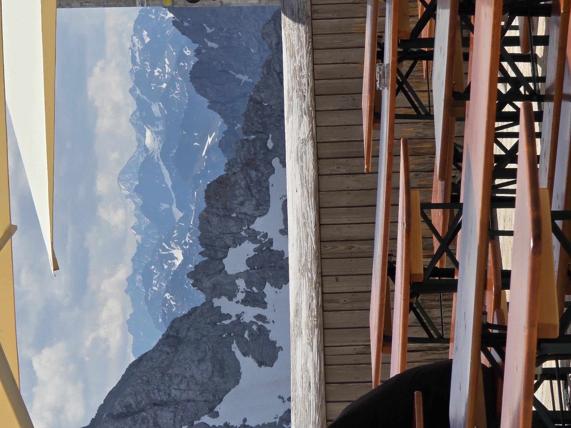 The image depicts an outdoor seating area with several wooden tables and benches, possibly at a restaurant or beer garden, overlooking a majestic mountain range. The tables are arranged in rows, with some partially obscured by others. In the foreground, a person wearing black clothing is seated at one of the tables. The background features a dramatic view of snow-capped mountains under a partially cloudy sky. The architecture suggests a location in a mountainous region, consistent with Garmisch-Partenkirchen, Germany, which is renowned for its alpine scenery and outdoor recreation facilities. The scene conveys a sense of tranquility and natural beauty, inviting visitors to relax and enjoy the panoramic views.
