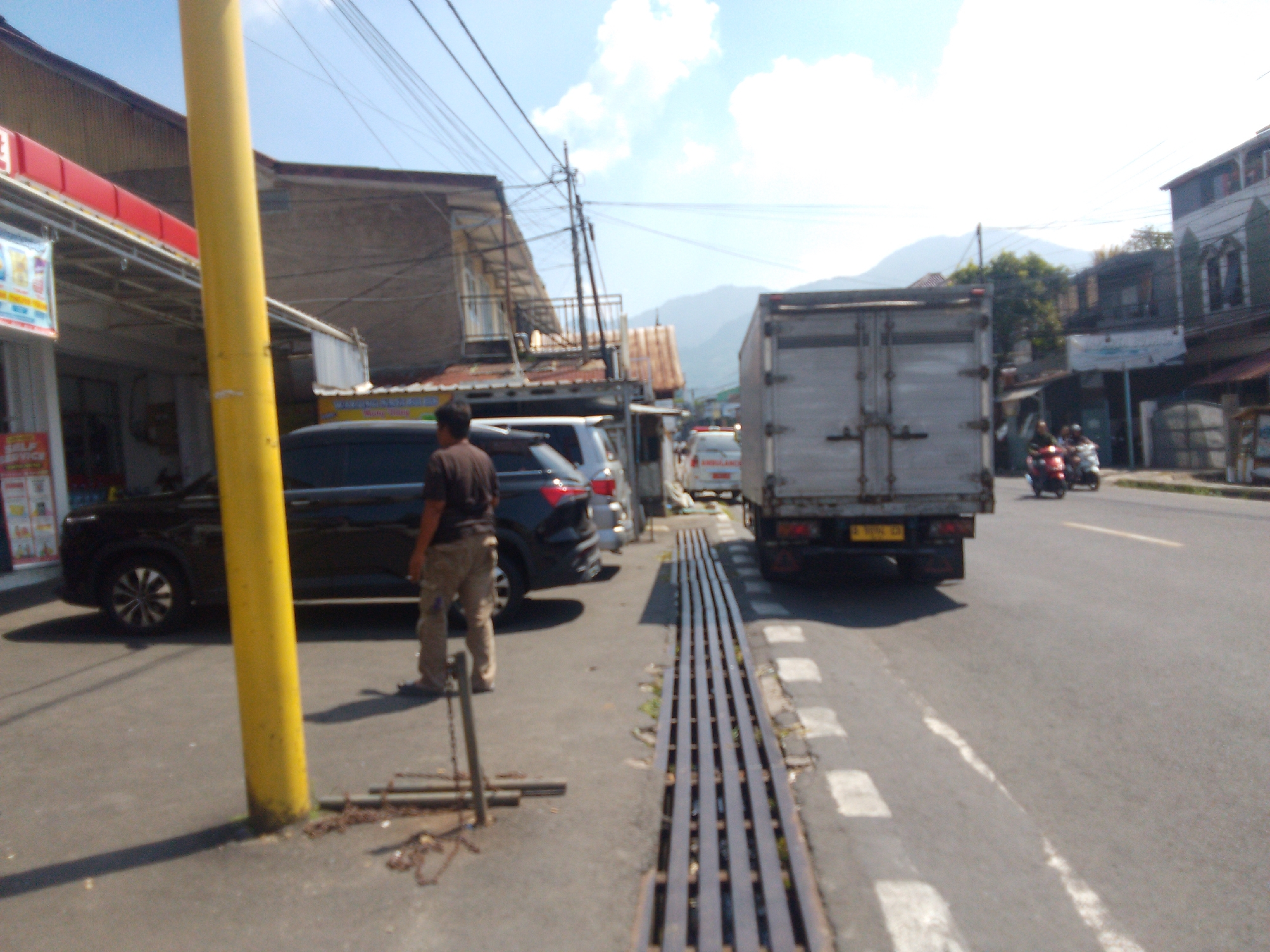 A man stands next to a dark colored sedan on a street in Cianjur, Indonesia. The man is wearing a dark shirt and light-colored pants, and is facing away from the camera. The sedan is parked on the left side of the image. A yellow pole is located in the foreground, obscuring part of the sedan. A large metal truck is driving on the right side of the street, heading away from the camera. The street appears to be lined with buildings and trees. The sky is blue with some clouds.