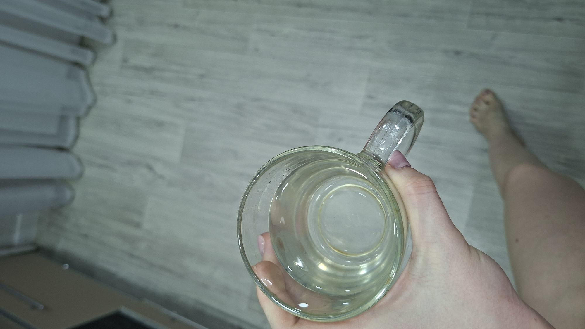 A top-down shot shows a hand holding a clear glass mug filled with a clear liquid. A bare foot and leg are in the background to the right. The floor is light gray with a wood-grain pattern. To the left, a white object with linear features is partially visible. Based on the visual information, it is impossible to determine if this image was taken in Chaikovsky, Russia.
