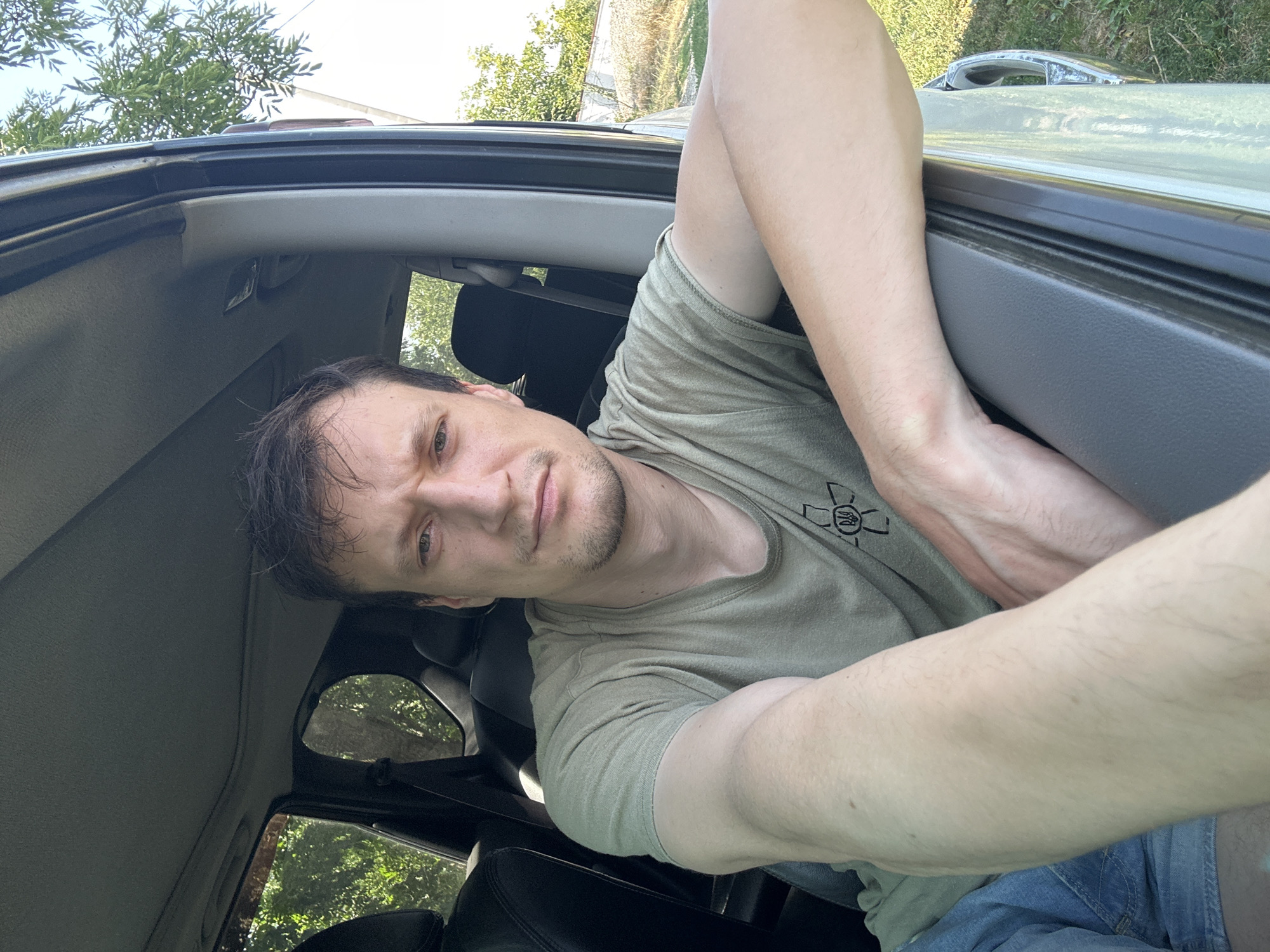 A man is photographed from the chest up, inside a vehicle, with his right arm resting on the open car door. He is wearing a light-colored t-shirt with a dark emblem on the left side of his chest. His left arm is bent at the elbow, with his hand resting on his chest. He has dark, medium-length hair and appears to be looking at the camera with a neutral expression. The interior of the vehicle is visible behind him, including the headrests of the seats. The car door is open, revealing a bright outdoor environment with green trees and a blue sky. The image has the appearance of a selfie. The location is Makiv, Ukraine.