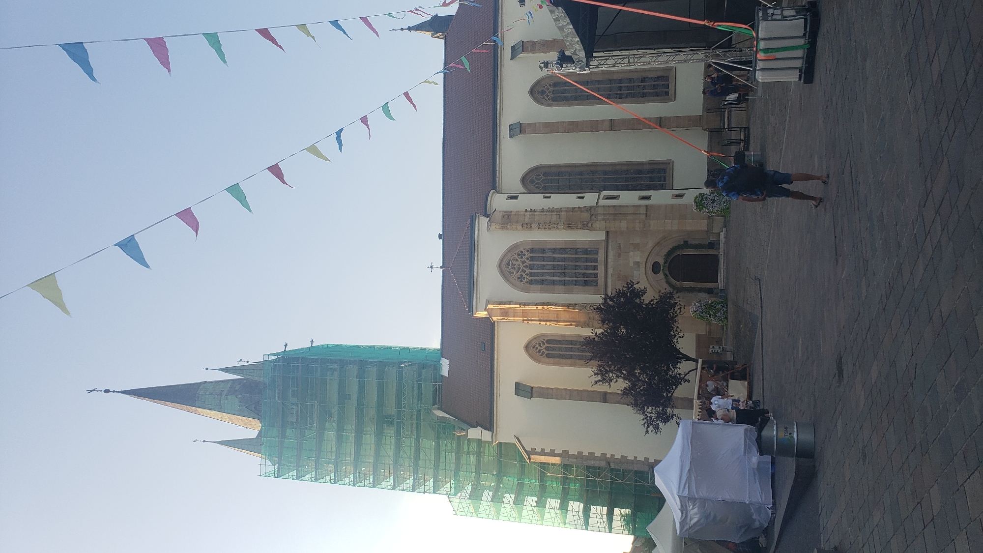 The image captures a view of the St. Nicholas Cathedral in Prešov, Slovakia. The cathedral's prominent spire is partially covered by green scaffolding, suggesting ongoing renovations. In the foreground, a man with a backpack walks away from the camera across a cobblestone plaza. Various tents and stalls are set up in the plaza, and a stage structure is visible on the right side of the image. Colorful triangular flags are strung across the sky, decorating the area. The sky is clear and bright, indicating a daytime scene, likely during fair weather.