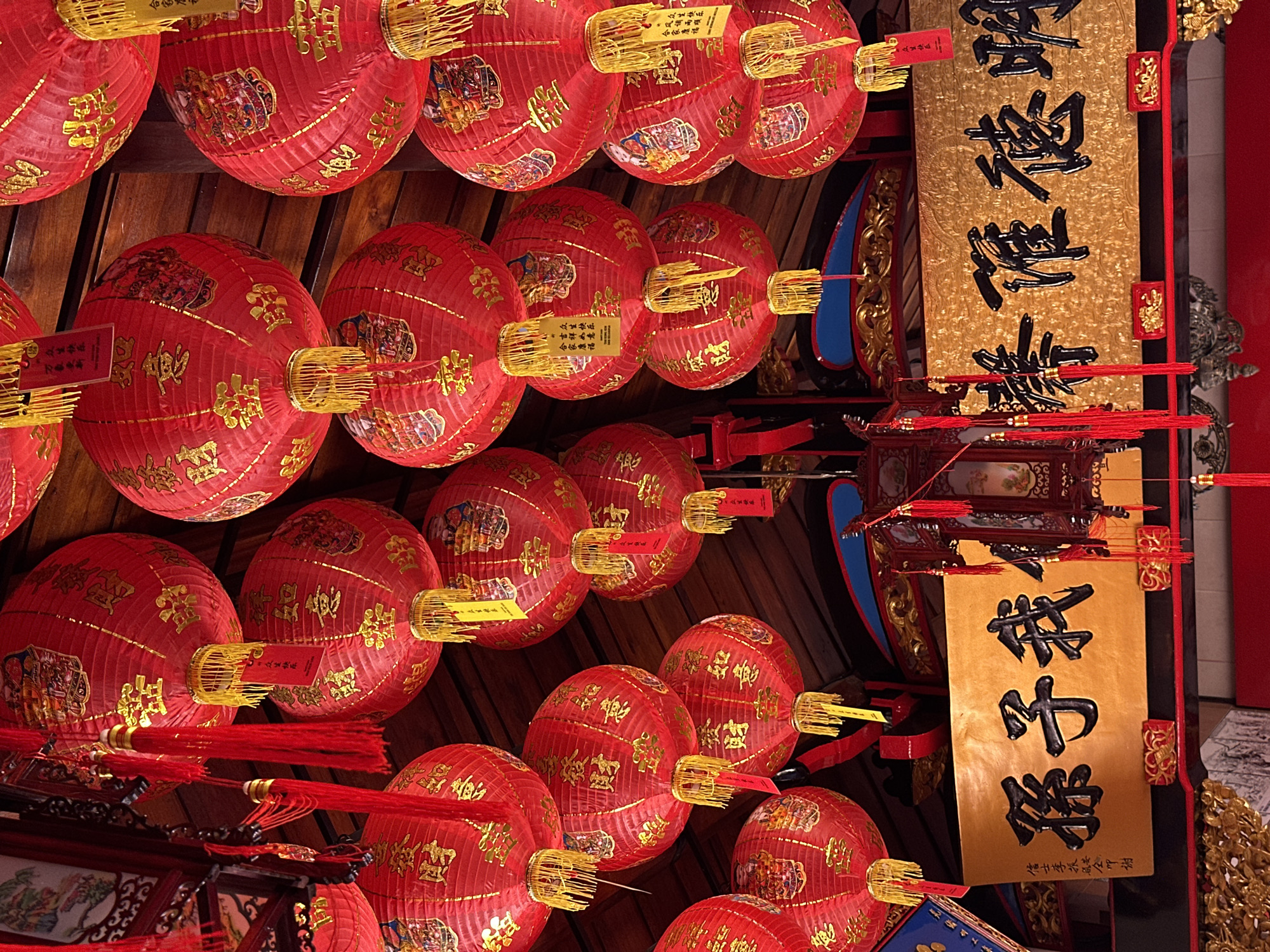 The image captures a detailed overhead view inside a building in Buleleng, Indonesia, characterized by numerous red Chinese lanterns suspended from a dark wooden ceiling. The round lanterns are adorned with gold Chinese characters, primarily "招財進寶" (ushering in wealth and prosperity) and "如意" (good fortune), and feature gold tassels and decorative tags at their bases. Many lanterns also display colorful pictorial motifs on their surfaces. Below the array of lanterns, two prominent rectangular golden plaques with raised black Chinese characters are affixed. The left plaque reads "我子孫" (my descendants), accompanied by smaller vertical text, while the right plaque displays "明德惟馨" (bright virtue is esteemed). Additional ornate red and gold architectural elements and intricate hanging decorations are visible in the lower portion of the frame. No individuals are depicted in the scene.
