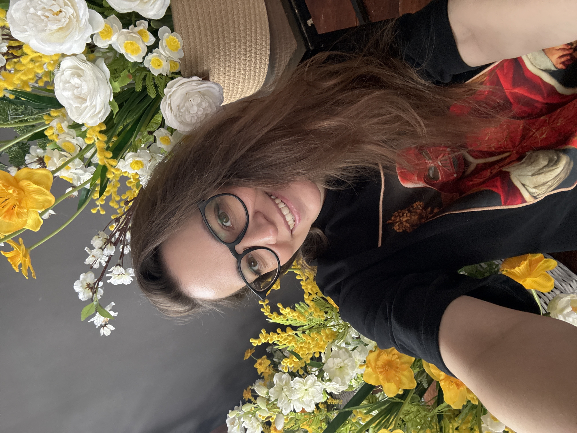 A person with shoulder-length brown hair and glasses smiles at the camera. They are wearing a black top with a red and white pattern on the shoulder. The person is surrounded by an abundance of white and yellow flowers. There is a straw hat partially visible in the background. The setting's location in Chaikovsky, Russia is not evident from the image.