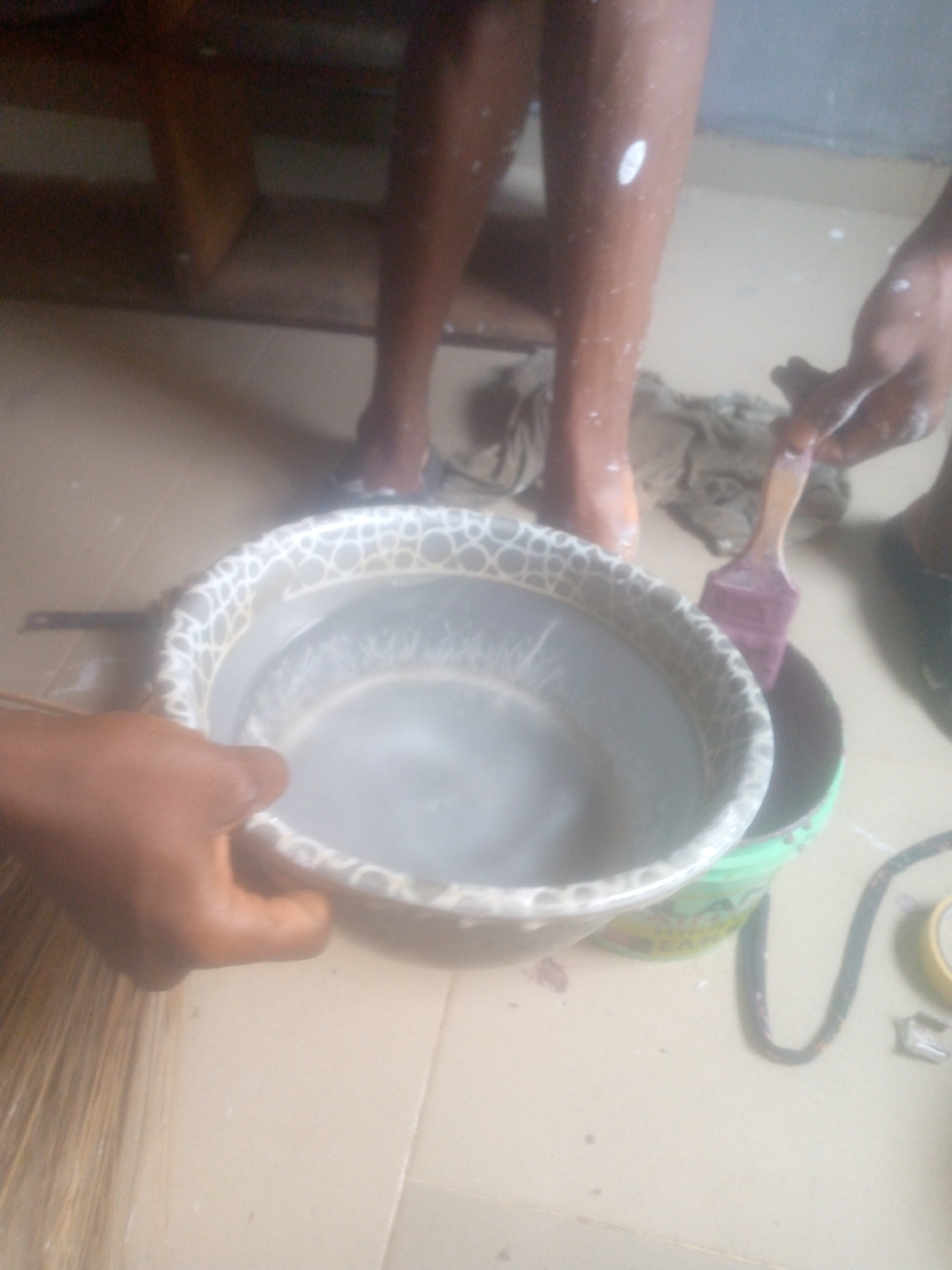The image captures an indoor scene, likely a workshop or a room undergoing maintenance. Two individuals are partially visible. In the foreground, a person's hand, with dark skin, holds a grey plastic basin patterned with white circles. The basin contains a cloudy, light-colored liquid, suggesting it's either water for washing or diluted paint. To the right of the basin, another person's hand, also with dark skin, holds a purple-bristled paintbrush, which is dipped into a green plastic bucket partially visible beside the basin.
In the background, another person's bare legs and feet are visible, notably covered with numerous white paint splatters, indicating their involvement in a painting activity. A light-colored, soiled cloth or rag is lying on the floor between their feet. The floor appears to be tiled or made of concrete, light beige in color. On the far left, a part of a broom-like object made of natural fibers is visible. To the right, a black electrical cord or hose is coiled on the floor, next to a small yellow object, possibly tape or a tool. The overall impression is one of active work, either in preparation for painting, during a painting task, or during the cleanup phase. No discernible text is visible in the image.