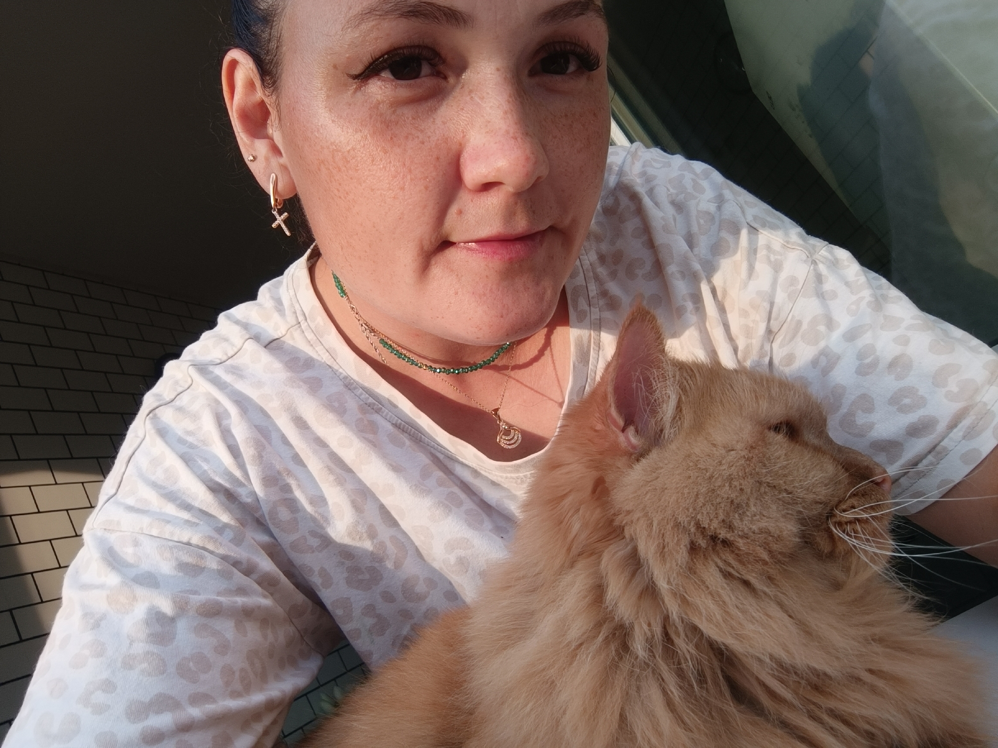 A woman is holding a fluffy orange cat. The woman has freckles, dark hair pulled back, and is wearing a white t-shirt with a light brown leopard print. She has on a cross earring and two necklaces, one with green beads and another with a delicate pendant. The cat is positioned on her chest, facing away from the camera. The background shows a tiled wall on the left and a reflection on the right, possibly from a window or glass surface. The lighting suggests it is either early morning or late afternoon. This image does not contain any recognizable landmarks or features that would allow for its identification in the city of Chaikovsky, Russia.