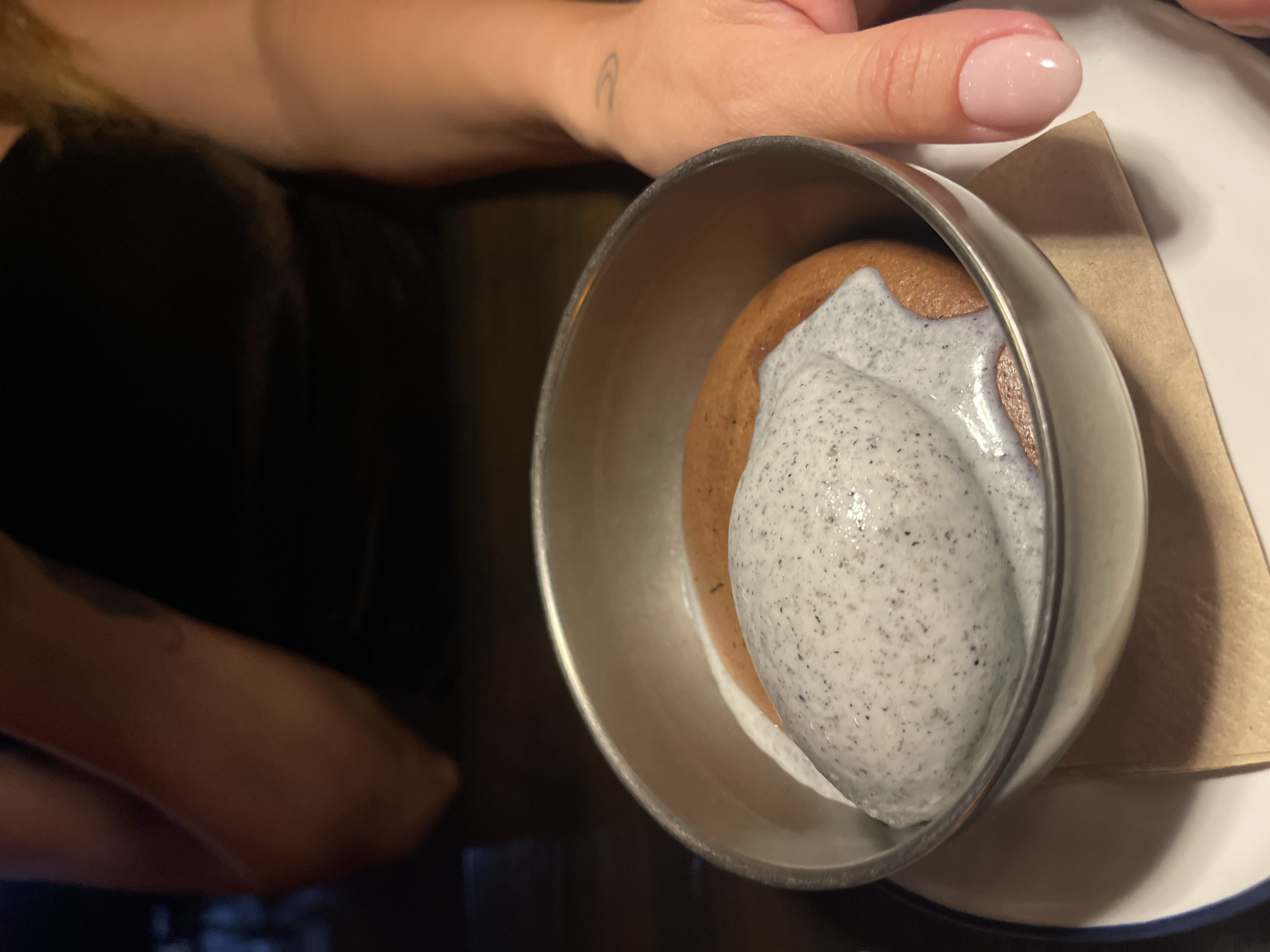 In a close-up shot, a serving of cookies and cream ice cream is presented in a metallic bowl. The ice cream is a pale grey speckled with dark cookie crumbs, and it sits atop a circular brown cookie. The bowl is placed on a white plate with a blue rim, and a folded brown napkin is tucked beneath it. In the upper portion of the image, a person's hand with light pink polished nails is visible, resting on the edge of the bowl. A faint tattoo of a small arch is visible on the person's forearm. The background is dark and slightly blurred, suggesting an indoor setting like a restaurant or cafe. The lighting is soft, highlighting the textures of the dessert and the skin of the hand. The overall mood is one of indulgence and enjoyment, likely a snapshot taken during a meal or dessert break.
