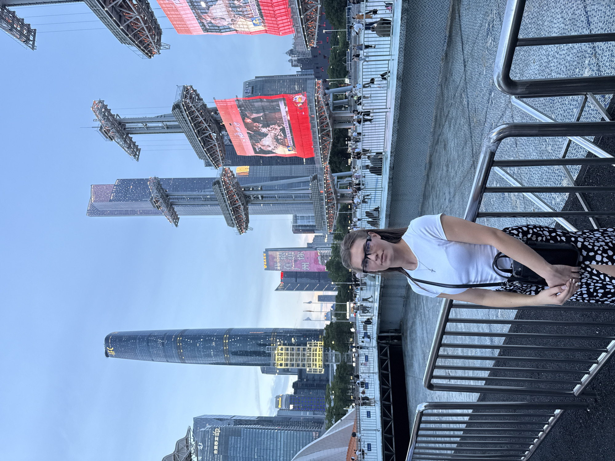 A young woman stands in front of a railing, looking at the camera. She is wearing a white t-shirt, a black and white patterned skirt, and glasses. A black purse is slung over her shoulder. Behind her, a modern cityscape is visible, with tall buildings and large screens displaying images and text. The sky is a dusky blue, suggesting it is either early morning or late evening. The scene appears to be in Tianhe District, China, likely near the Zhujiang River. A crowd of people can be seen in the background, gathered on a walkway or plaza.