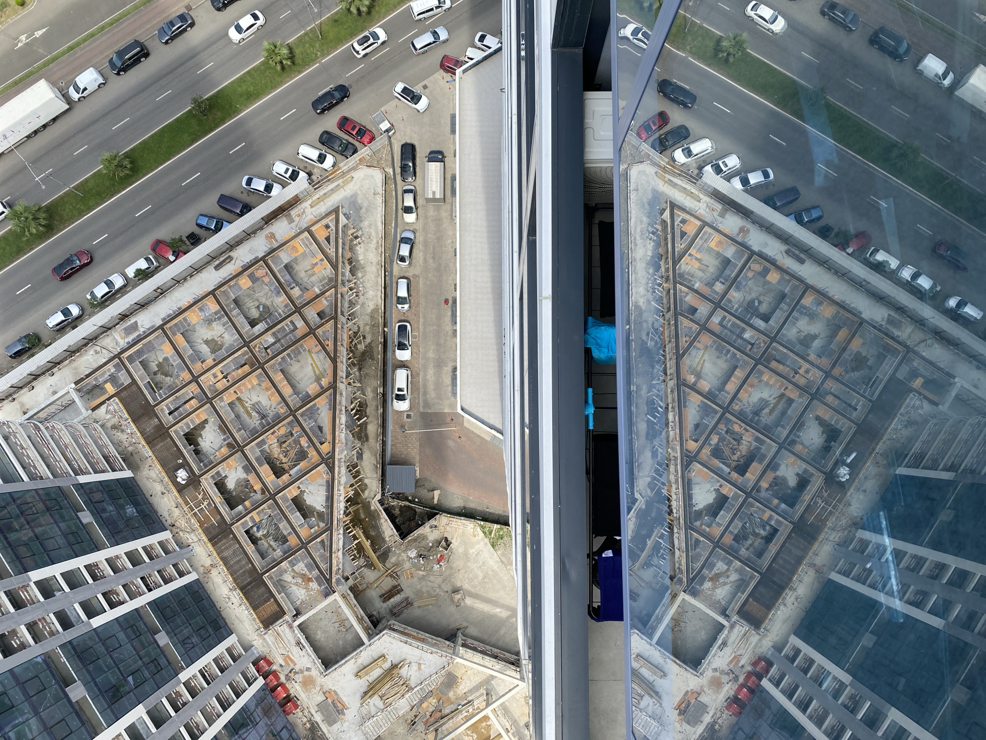 This is an aerial, high-angle, full-shot photograph of an urban construction site located in Batumi, Georgia. The scene is divided horizontally by a large building with mirrored glass windows, reflecting the construction site and a multi-lane road with parked vehicles and traffic.
The upper portion of the image shows the construction site. A partially completed multi-story building with a grey facade and dark glass windows is visible on the left. Adjacent to it is a large, open foundation area with a complex network of concrete pillars and rebar, forming triangular and rectangular sections. Wooden scaffolding and construction materials are scattered throughout this area. A paved road runs parallel to the construction site, lined with trees and grass. Numerous vehicles, including cars, trucks, and vans, are parked along the road and driving on it.
The lower portion of the image is dominated by the reflection in the glass of another building. This reflection reveals a similar construction site with concrete foundations and wooden structures, mirroring the elements seen in the upper part of the photograph. Below the mirrored building, a section of a road with parked cars is visible. A few palm trees are scattered throughout the scene.
The lighting suggests it is daytime, likely mid-morning or afternoon, with bright sunlight casting shadows. The overall impression is one of ongoing urban development and infrastructure.