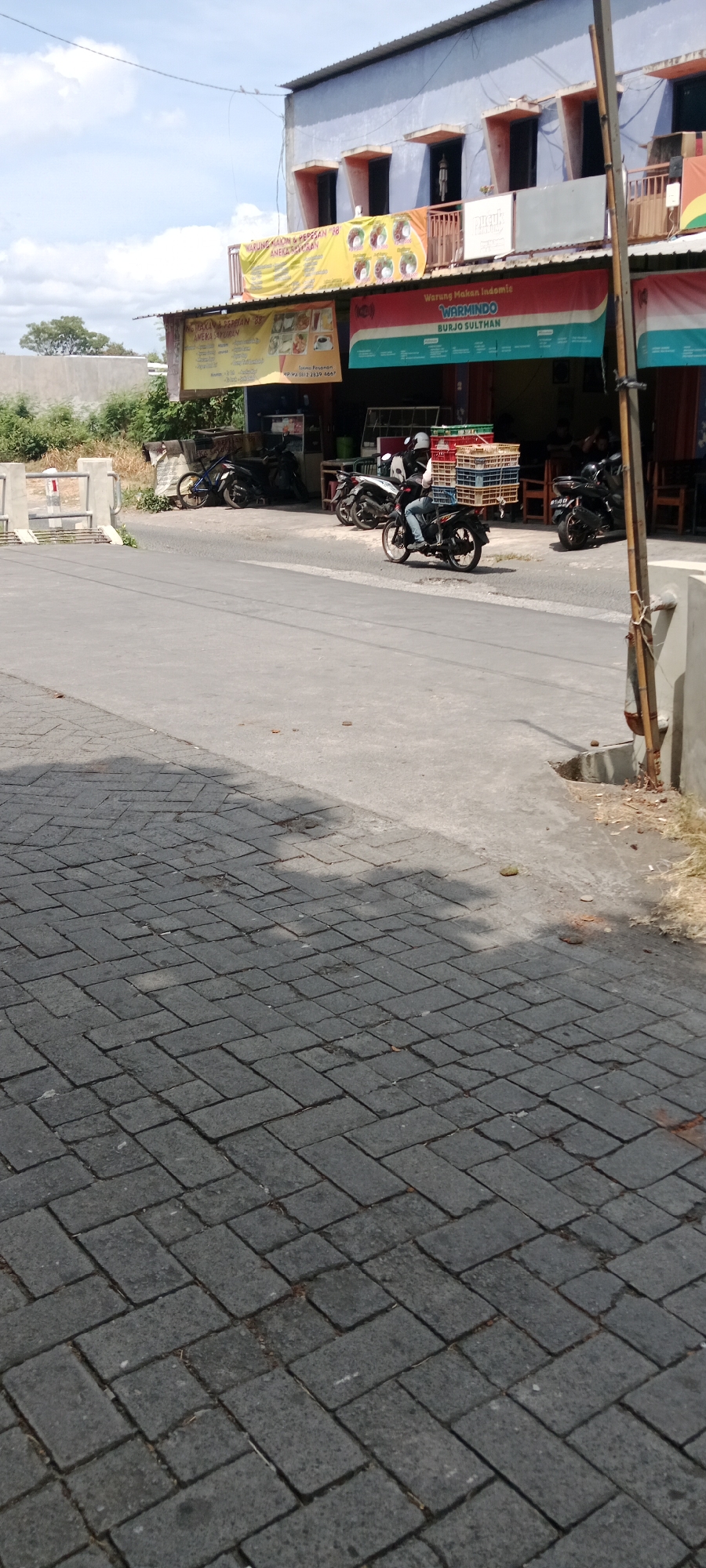 The image depicts a street scene in Maguwoharjo, Indonesia, with several shops lining the right side of the road. A motorcycle rider, wearing a helmet, is in the process of passing by, carrying a stack of red and blue crates on the back of the motorcycle. The crates appear to be made of plastic and are stacked several layers high. Parked motorcycles are visible in front of the shops, along with tables and chairs, suggesting an outdoor seating area. Banners advertising food establishments are displayed on the buildings. The road is paved with asphalt, and the foreground shows a pedestrian walkway made of interlocking grey bricks. The sky is bright with some clouds, indicating it is daytime. The overall impression is of a typical commercial street in an Indonesian town.