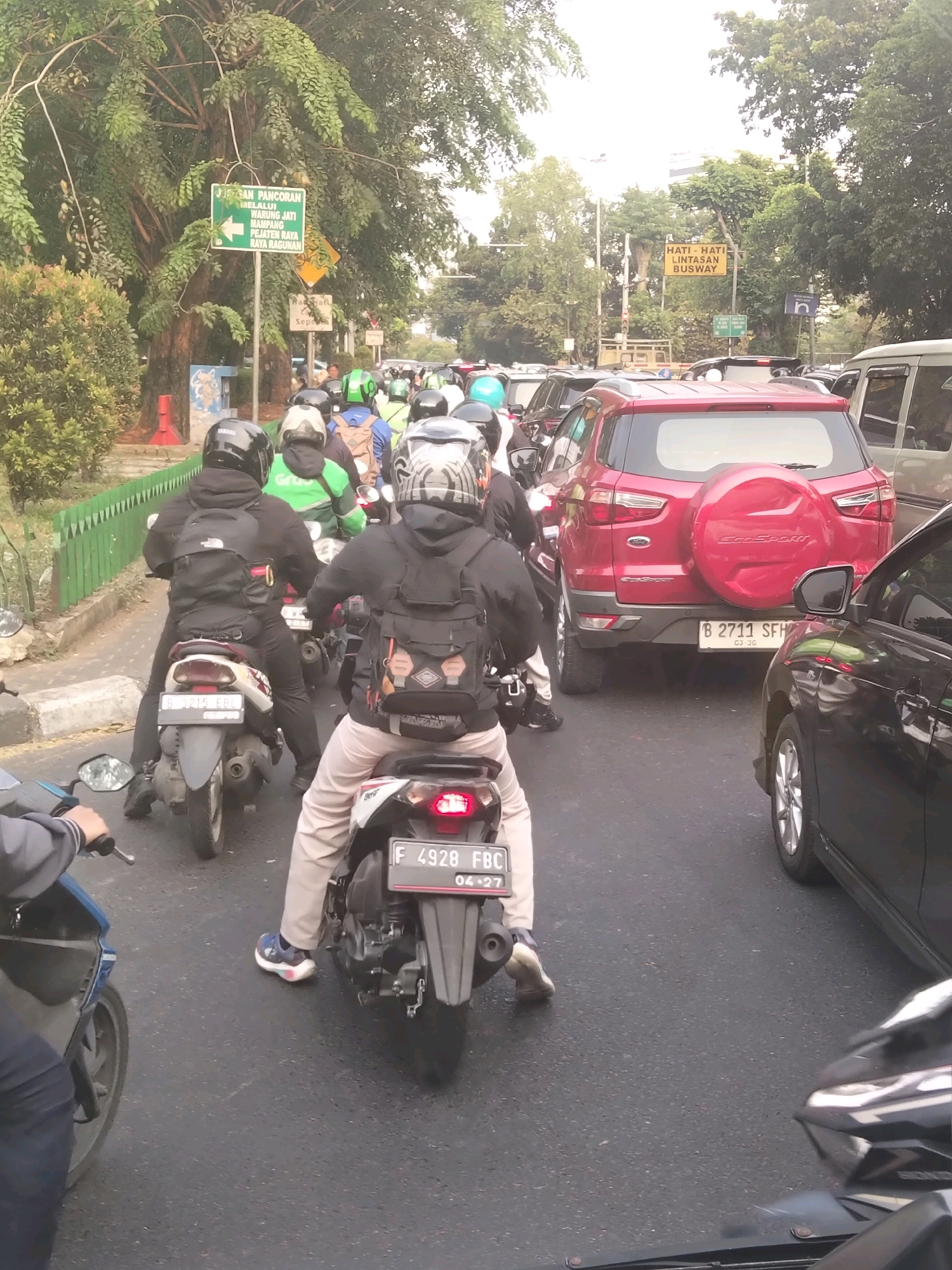The image depicts a congested street scene in South Jakarta, Indonesia, likely during daytime with overcast conditions. The foreground features several motorcycles and a portion of a blue scooter. The riders are wearing helmets, with one rider in the center wearing a black jacket and a backpack, and another on a white and red motorcycle with a black backpack. Behind these, a red SUV is visible, followed by other vehicles and more motorcycles. Traffic appears to be at a standstill or moving very slowly. Signage can be seen in the background, including a green sign with Indonesian text and an arrow, and a yellow sign with the text "HATI - HATI LINTASAN BUSWAY" which translates to "BEWARE BUSWAY CROSSING." This suggests the location is near a busway corridor, a common feature in Jakarta's transportation infrastructure. The environment includes trees lining the street, a green fence on the left, and a paved road. The overall impression is one of urban traffic congestion typical of a major city.