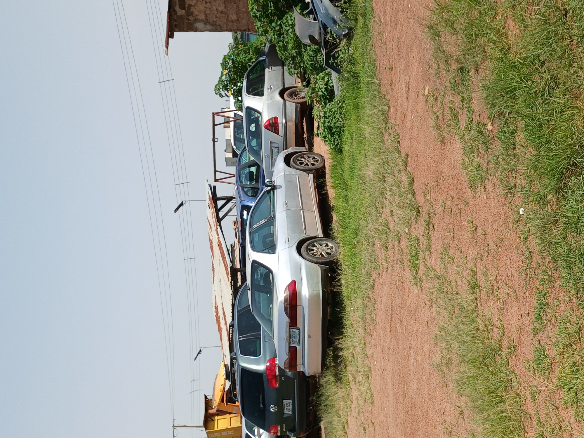 The image depicts a car junkyard in Gura Top, Nigeria. In the foreground, a silver sedan is parked on a dirt and grass area, its rear bumper facing the camera. To its left, a dark green Volkswagen Touareg SUV is parked, also with its rear facing the camera. Behind these vehicles, various other cars are visible, some partially obscured by foliage. A yellow construction vehicle is parked to the left of the frame, and a partially dilapidated corrugated metal roof structure stands in the background. The sky is clear and pale blue. The scene appears to be an outdoor storage or disposal area for vehicles.