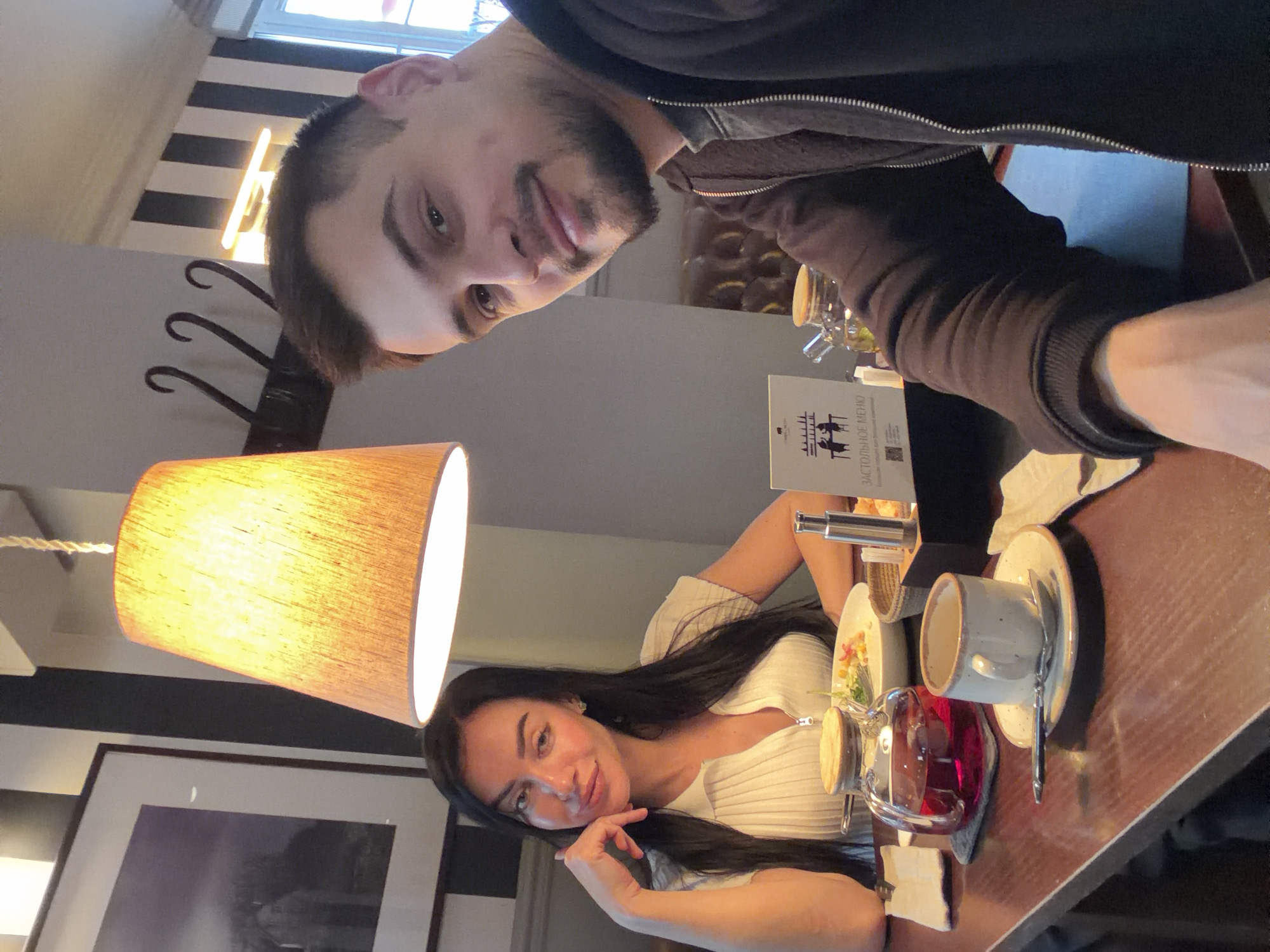 Two people are sitting at a table in a cafe. A man with short dark hair and a beard is facing the camera. A woman with long dark hair is sitting across from him. The table has a cup and saucer, a pot of tea, and a plate of food. A lamp is hanging over the table. The cafe is in Krasnoyarsk, Russia.