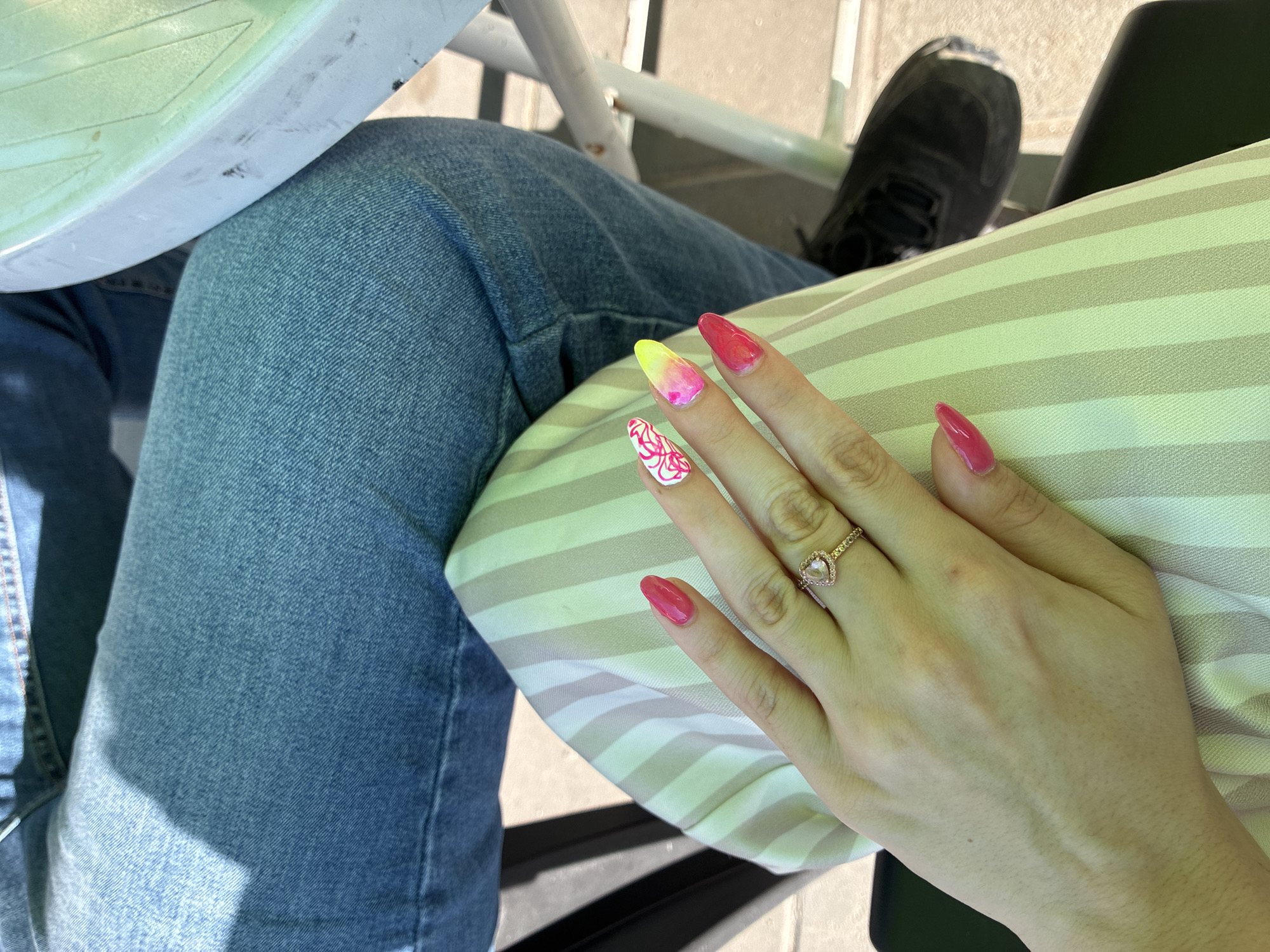 A close-up shot captures a woman's hand with a pink heart-shaped ring and brightly colored, long, almond-shaped nails, adorned with pink, yellow, and white abstract designs. The hand rests on a light green and cream striped fabric, likely part of a dress or skirt. In the background, a person wearing blue denim jeans is visible, along with parts of chairs and a table, suggesting an outdoor seating area. The lighting indicates a sunny day. This scene is likely in Aglen, Bulgaria, based on the provided location.