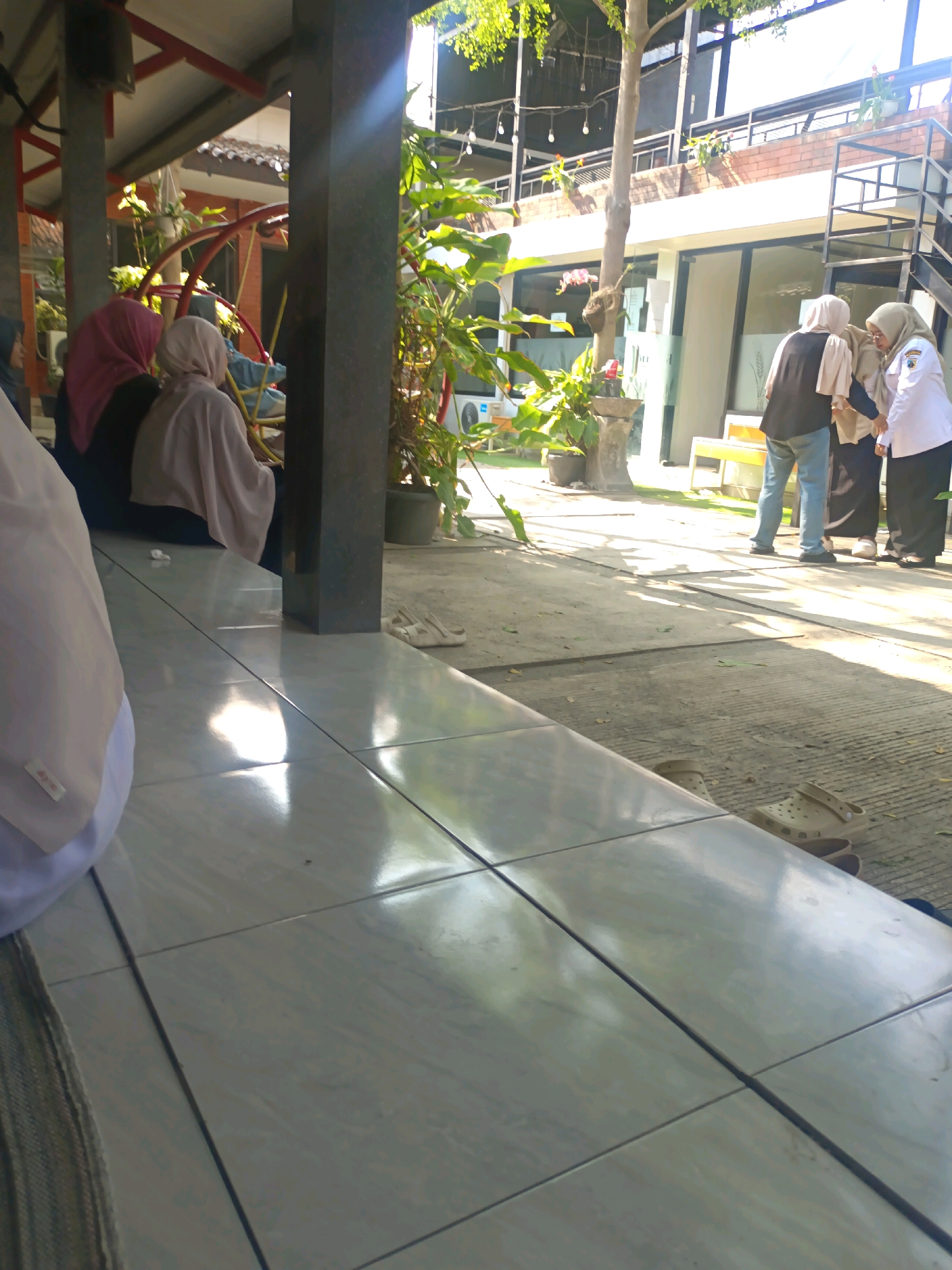 The image shows a tiled seating area with a view of a modern building and outdoor seating. In the foreground, a person in a white garment, likely a hijab or abaya, is partially visible from the side. To the left, two individuals are seated, both wearing hijabs, one in pink and the other in light grey. Behind them, a yellow circular structure is visible, possibly part of a playground or decorative element. In the mid-ground, three people are interacting. One person in a dark vest and jeans is facing away, speaking to two individuals in hijabs. One of these individuals is wearing a light-colored hijab and a dark outfit, while the other is in a white uniform with a dark skirt and a light-colored hijab, possibly a medical professional or uniform. The background reveals a multi-story building with large windows and balconies. The environment appears to be an outdoor cafe or public space. The lighting suggests it is daytime, with strong sunlight casting shadows. The location is identified as Kerangkulon, Indonesia.