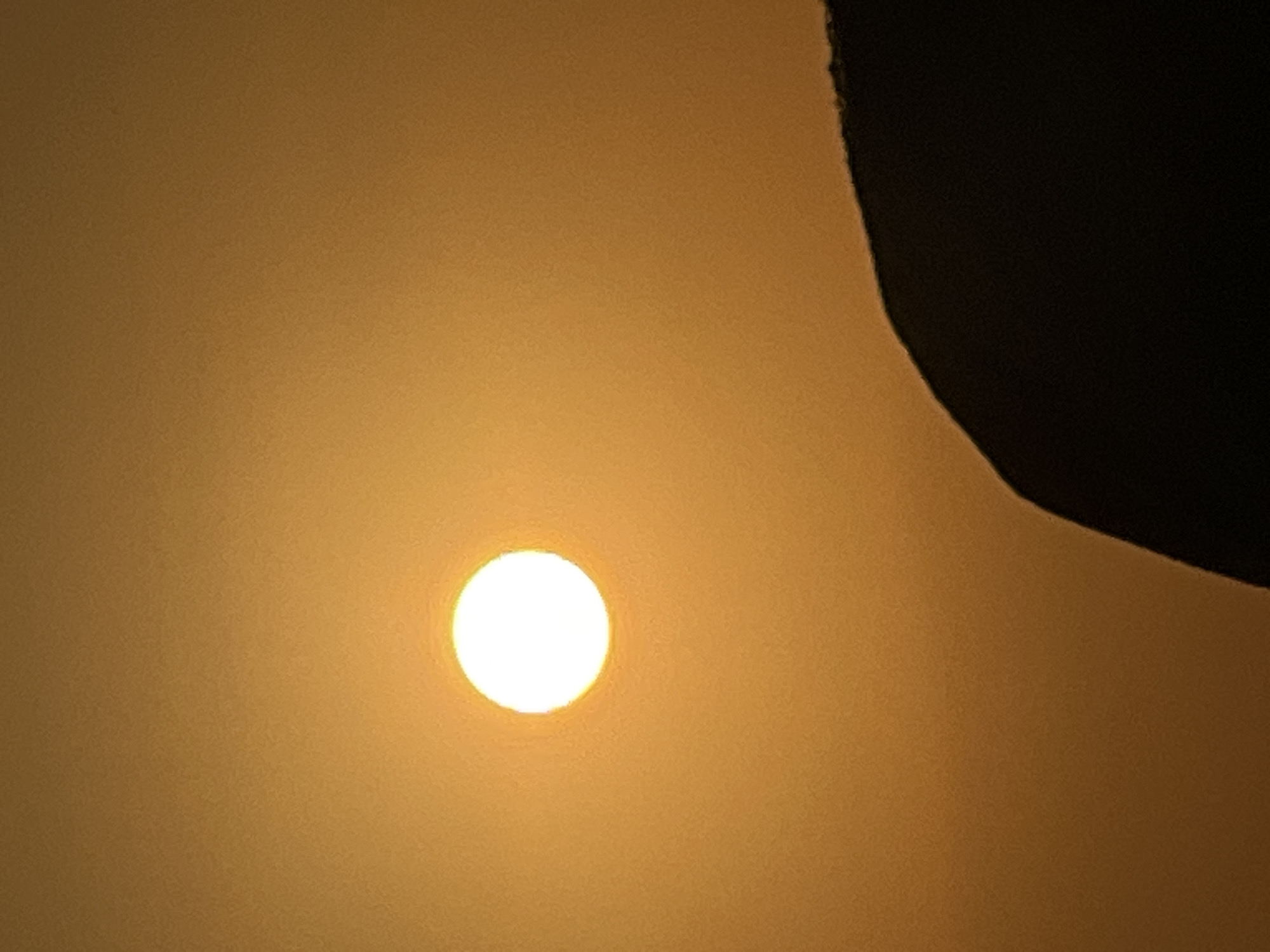The image shows a bright, circular sun against a hazy orange sky. In the lower right portion of the frame, a dark, silhouetted object, possibly a hill or a building, curves into the frame. The sun is positioned in the upper left quadrant of the image. The overall lighting suggests either sunrise or sunset. There are no discernible people, animals, or specific landmarks that would indicate the location in Kuta, Indonesia, beyond the assumption provided in the prompt. The scene is devoid of any discernible activity or interaction.
