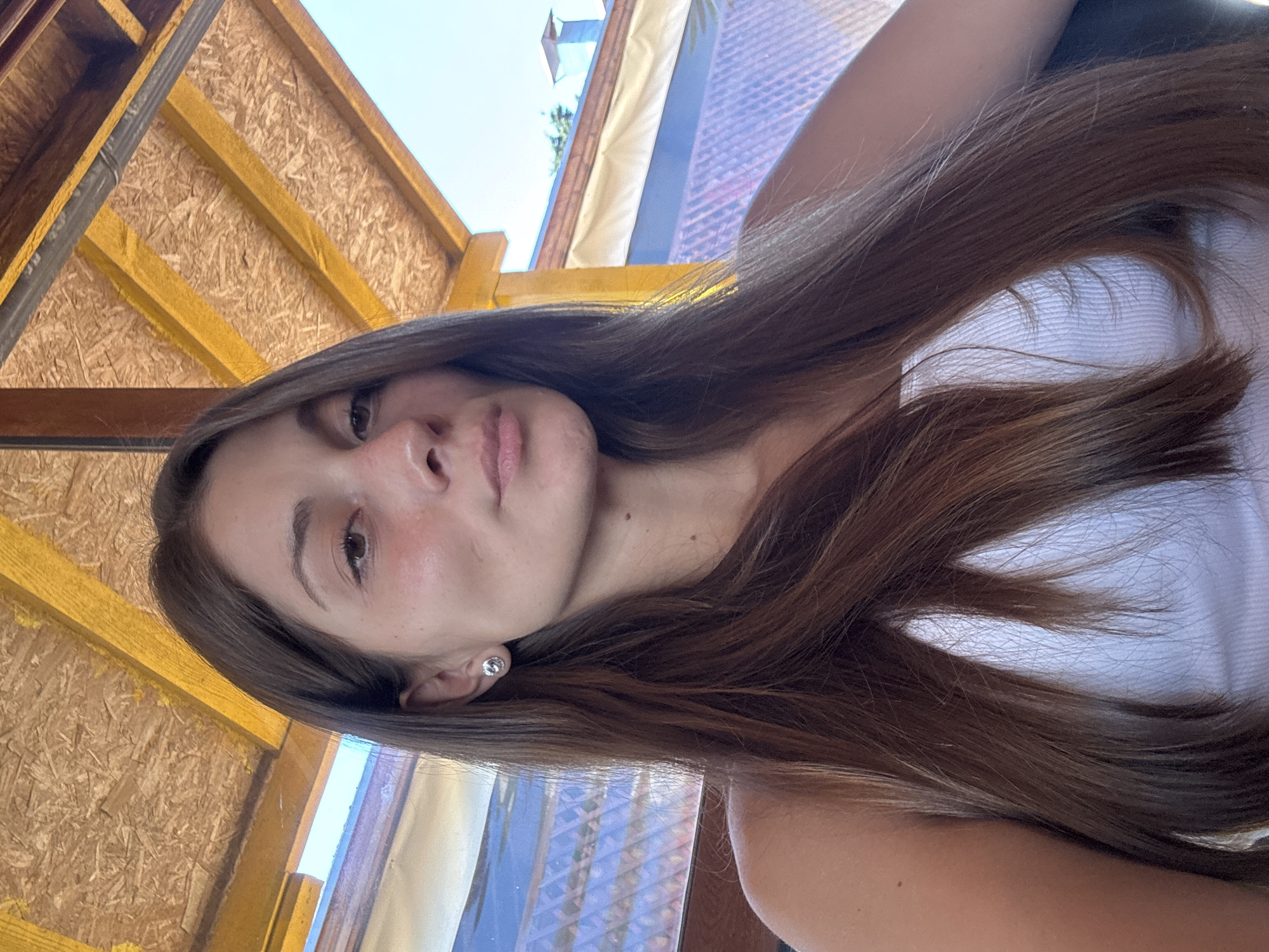 A young woman with long, brown hair is the sole subject in this portrait. She is captured in a close-up, vertical orientation, with her face angled towards the camera. Her expression is neutral, with her gaze directed slightly away from the lens. She is wearing a simple white tank top, and a small, sparkling stud earring is visible on her left ear.
The background suggests an outdoor setting, possibly a covered patio or balcony. Wooden beams, typical of construction or roofing, form a textured backdrop. Beyond these, a glimpse of a blue sky and a patterned, translucent covering can be seen, hinting at a sunny day and perhaps a warm climate. The overall lighting suggests it is daytime. The image seems to be a selfie, taken by the subject herself.
The scene has a casual, relaxed feel, possibly indicating a moment of downtime or a brief pause during an outdoor activity. There is no visible text in the image. The location context provided identifies Sozopol, Bulgaria, as the setting, which aligns with the outdoor, potentially coastal, vibe suggested by the image.