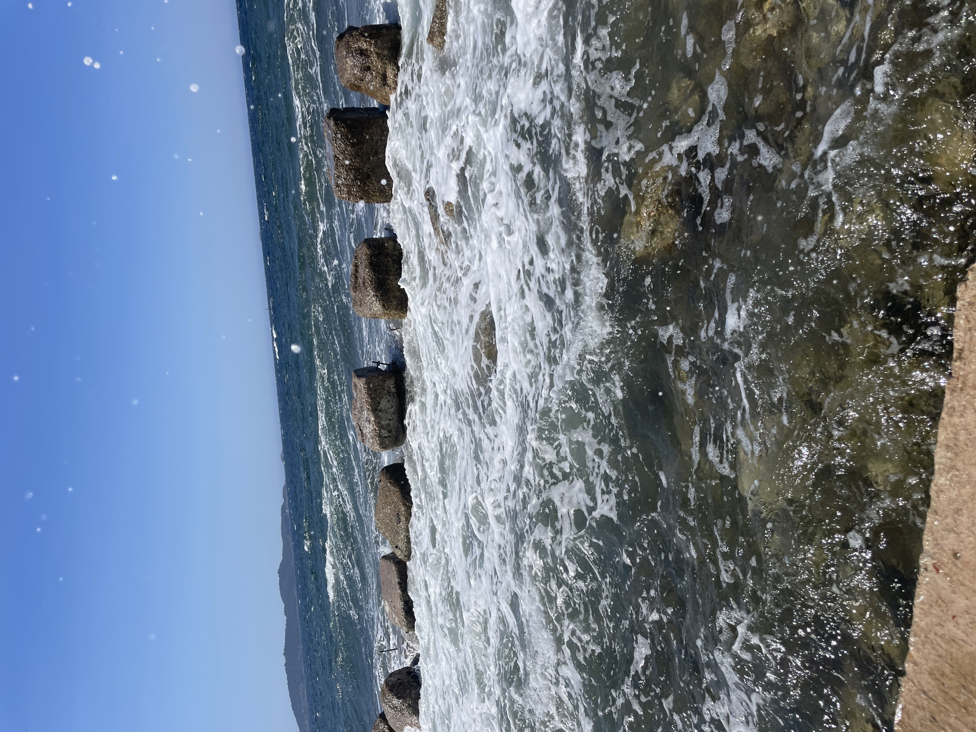 The image shows a coastline in Carqueiranne, France, under a clear blue sky. Large, rough-hewn stones, likely part of a breakwater or pier, are partially submerged in the sea. Waves are crashing against these stones, creating white foam and turbulent water. The sea is a deep blue, with a distant island or landmass visible on the horizon. The foreground features shallow water with visible rocks and a concrete or stone ledge. There are no people visible in the image.