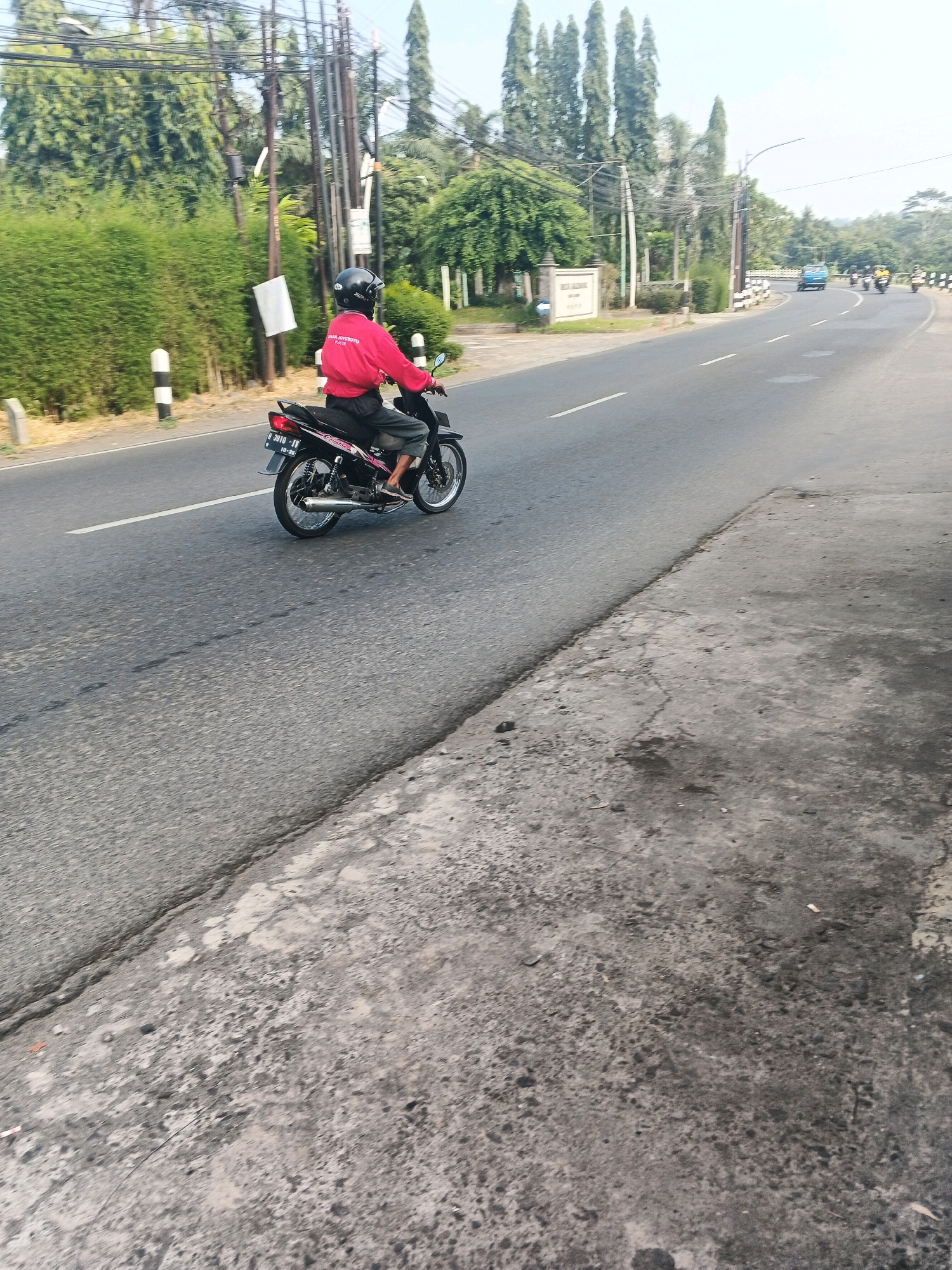 A lone motorcycle rider, wearing a red jacket and black helmet, is traveling on a paved road in Ungaran, Indonesia. The rider is positioned in the left lane, moving away from the camera. The motorcycle is black with silver accents. The road is bordered by a low, manicured green hedge on the left and a concrete shoulder on the right. In the background, there are tall trees, utility poles with wires, and a signpost. Further down the road, other vehicles, including a blue van and at least two other motorcycles, are visible in the distance. The sky is clear and bright, suggesting a sunny day.