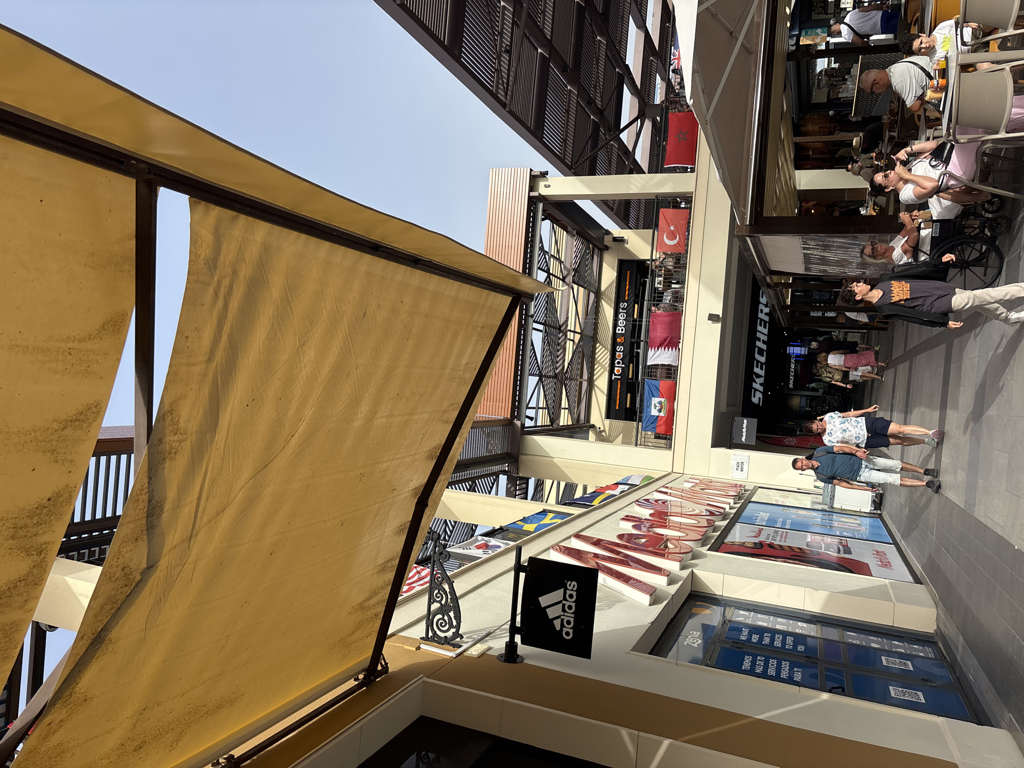 The image depicts an outdoor pedestrian walkway lined with shops and eateries under a covered awning. The location is identified as Orihuela, Spain, specifically within an area featuring flags of various countries displayed on balconies. A prominent Adidas store sign is visible on the left, with a large, red-lettered sign for "Mercado de..." (likely Mercado de Abastos, a common name for markets in Spain) above a storefront with blue doors and informational posters. Further down the walkway, a Skechers store is identified. People are seen walking and seated at outdoor tables of a cafe. The atmosphere appears to be a commercial area, possibly a shopping center or a street with retail establishments. The architectural style features exposed wooden beams supporting a yellow fabric awning and segmented roof structures. The sky is clear and blue.