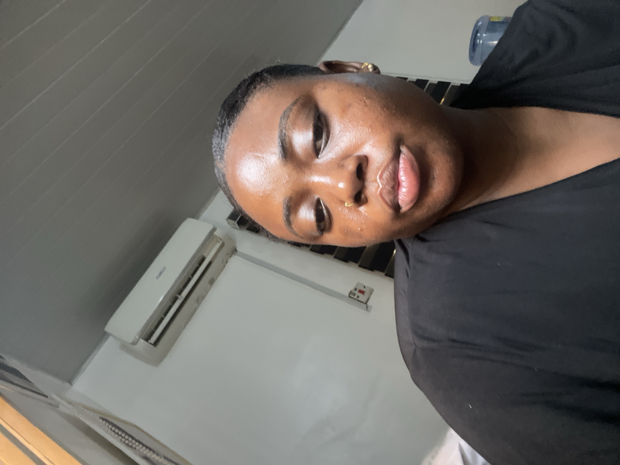 A close-up photograph shows a person with dark skin and short, dark hair. The person is wearing a black top and has piercings in their ear and nose. The person's skin appears somewhat shiny, possibly due to sweat or oil.
The background includes an air conditioning unit mounted on a white wall. The ceiling is white and appears to be paneled. There are window blinds visible along the left edge of the image.
There is not enough information to determine a specific location within Abuja, Nigeria.
