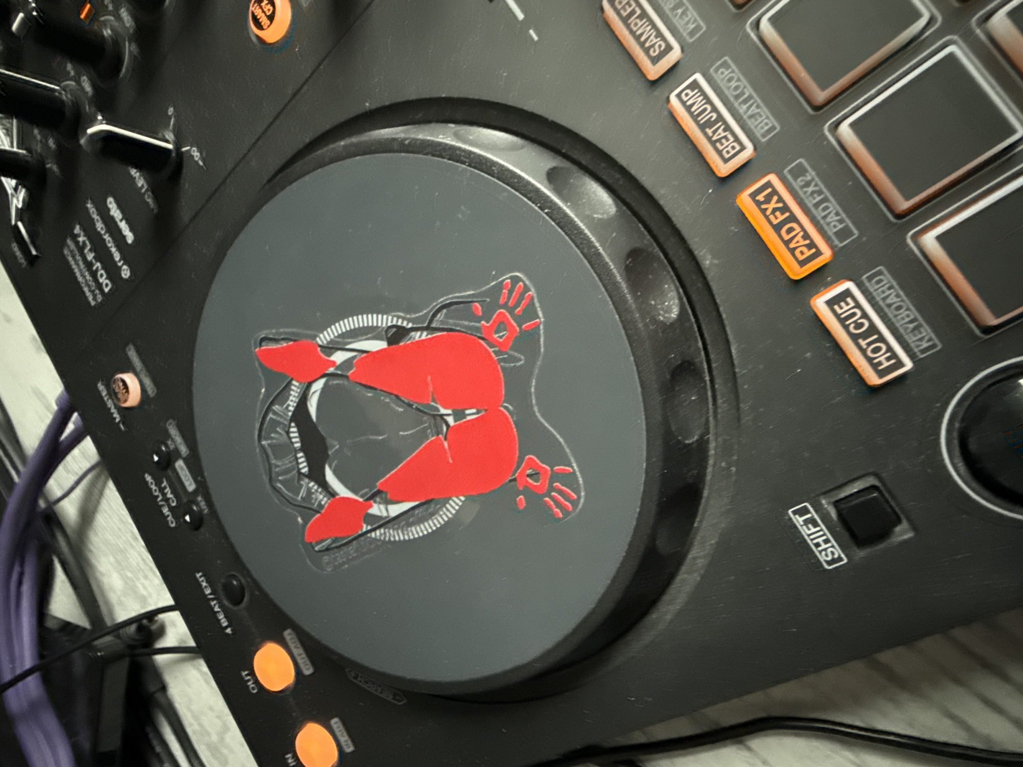 The image shows a close-up of a black Pioneer DDJ-FLX4 DJ controller. A sticker depicting a red anatomical rendering of a human buttocks with hands is affixed to the left jog wheel. Control buttons and labeled pads are visible to the right of the jog wheel, including "HOT CUE," "PAD FX1," "BEAT JUMP," and "SAMPLE." The "SHIFT" button is also visible. Other labeled buttons and features are visible on the periphery of the image. Purple cables are partially visible on the left side of the image. The location of the image is not specified.