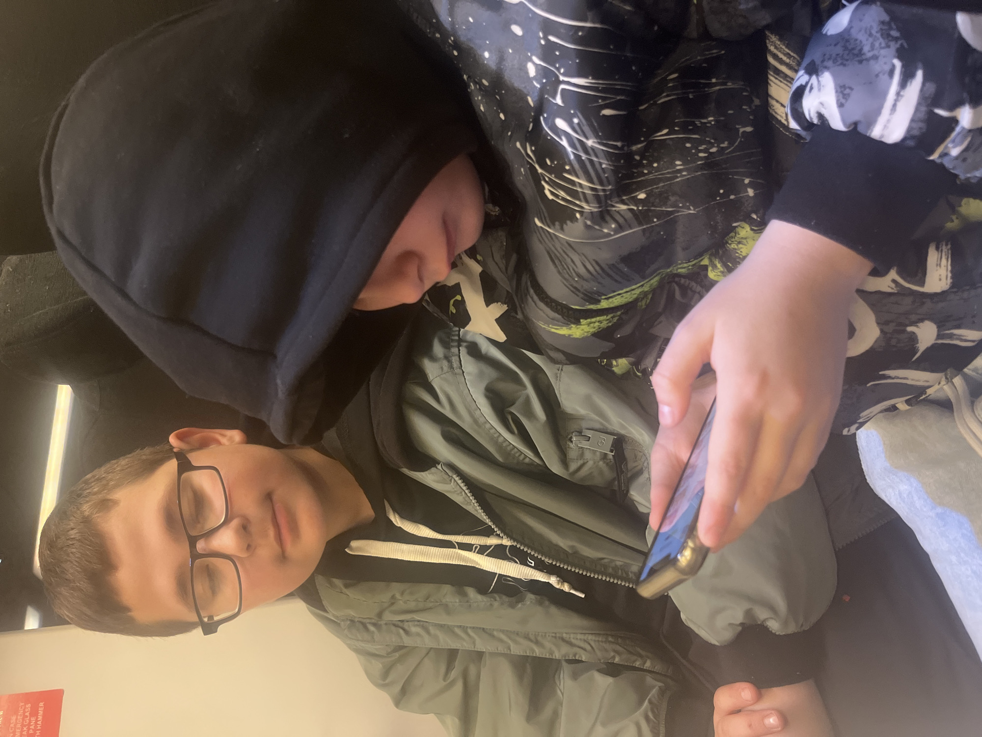 Two children sit next to each other. The child on the left wears glasses and an olive green jacket with white drawstrings. The child on the right wears a dark hooded sweatshirt with white and lime green splashes. The child on the right is holding a cell phone. It is unknown whether the photo was taken in Sochi, Russia.
