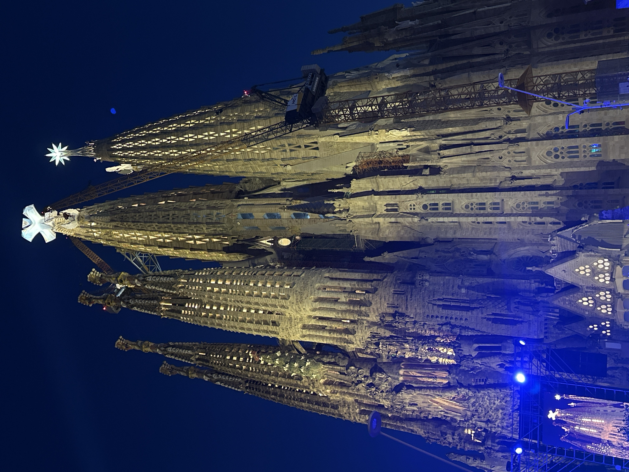 The image shows a nighttime view of the Sagrada Familia basilica in Barcelona, Spain. The basilica is illuminated with warm yellow lights, highlighting its intricate Gothic architecture. Several tall towers rise towards a deep blue night sky, topped with decorative spires and illuminated crosses. Construction cranes are visible, indicating ongoing work on the structure. In the foreground, there are blue stage lights and scaffolding. The overall scene is majestic and awe-inspiring, showcasing the ongoing construction and illumination of this iconic landmark.