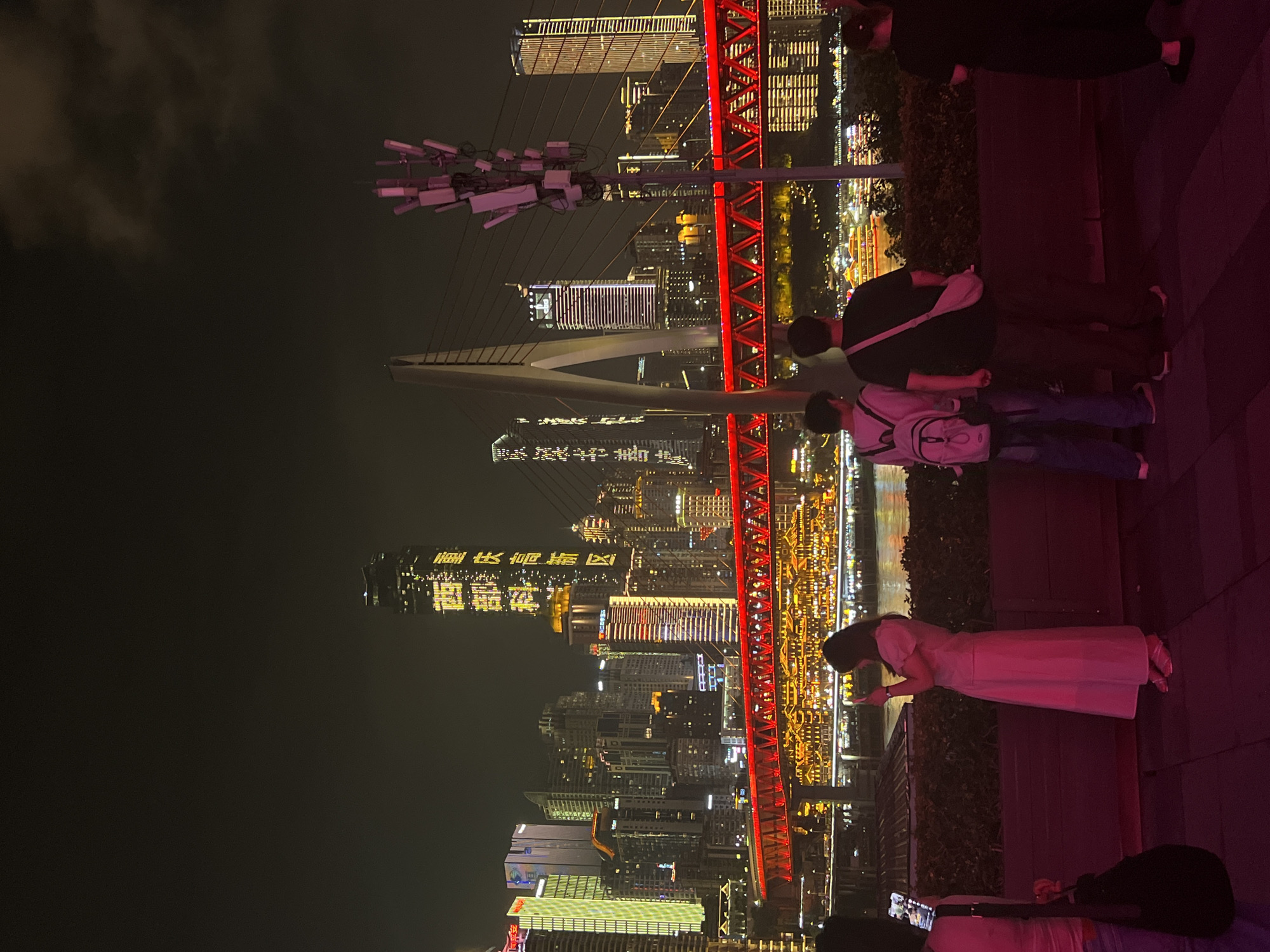 A night view of the Yangtze River in Jiangbeicheng Subdistrict, Chongqing, China. A city skyline with numerous illuminated skyscrapers is visible in the background, with a red illuminated bridge spanning across the river in the mid-ground. Several people are standing on a viewing platform in the foreground, facing the city. One woman in a pink dress is holding a phone and taking a picture. Two men are standing together, one with a white backpack. Another person is standing on the far right. The river is lined with glowing lights from buildings and boats. A cell tower with pink lights is positioned to the right of a suspension bridge pylon. The sky is dark with some clouds.