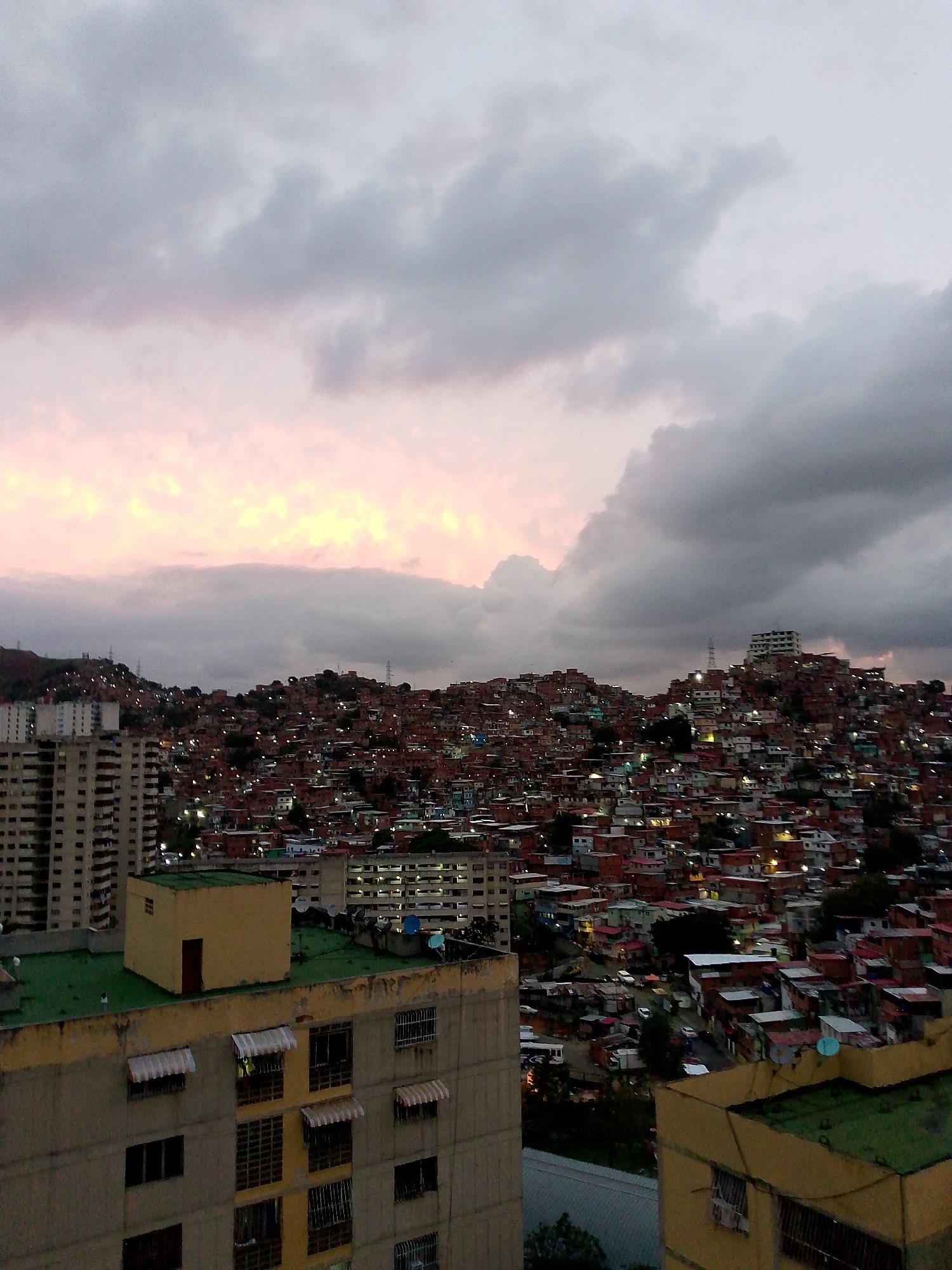 An overcast evening scene shows a densely populated hillside in Caracas, Venezuela. A cluster of closely packed, mostly brick and white structures covers the slope, interspersed with a few larger buildings and illuminated by numerous scattered lights. In the foreground, the roofs of apartment buildings are visible, with glimpses of building facades and windows. The sky is filled with layered clouds, tinted with muted pink and gray hues.