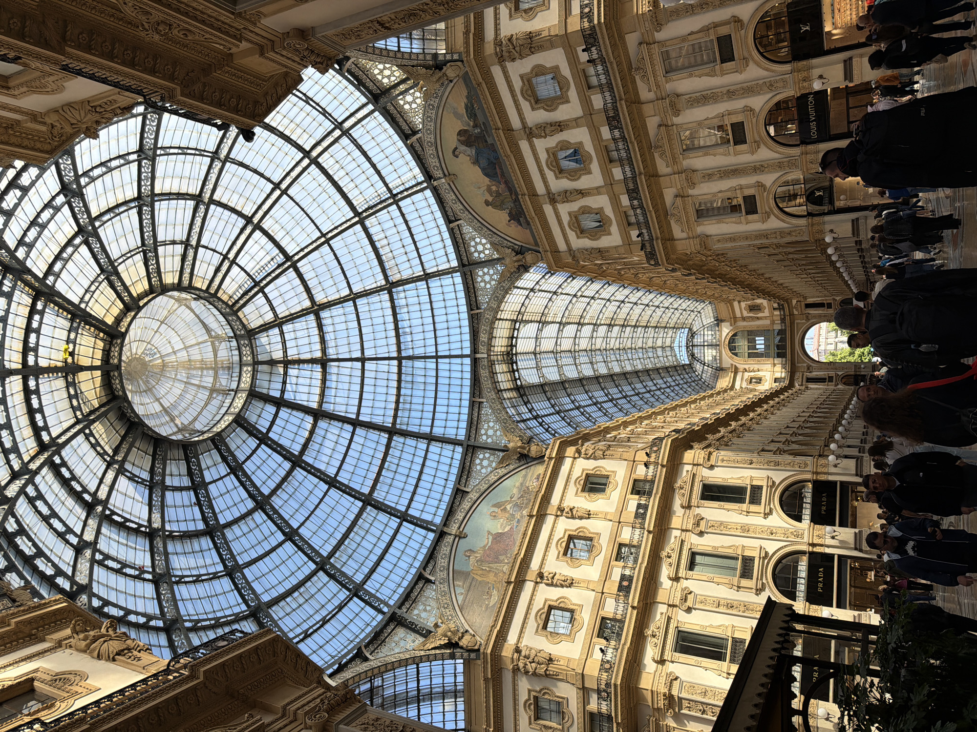 This image captures the interior of a grand, ornate building, which is unmistakably the Galleria Vittorio Emanuele II in Milan, Italy. The dominant feature is the magnificent glass and iron-framed dome and barrel-vaulted ceiling, allowing abundant natural light to stream in from a clear blue sky, indicating a sunny daytime.
The architecture is opulent, featuring multi-story buildings lining the gallery with intricate carvings, decorative friezes, and balconies. Arched windows and elaborate decorative elements adorn the facade. Several people are visible throughout the gallery, walking along the tiled floor. They appear to be a mix of shoppers and sightseers, creating a bustling, lively atmosphere typical of a public attraction or shopping destination.
Notable luxury brand storefronts line the passage. Clearly visible text on the shop signs includes "LOUIS VUITTON" and "PRADA," confirming the high-end retail environment. The perspective looks upwards and along the extensive length of the gallery, emphasizing the impressive scale and intricate details of the ironwork and glass roof, as well as the richly decorated walls.