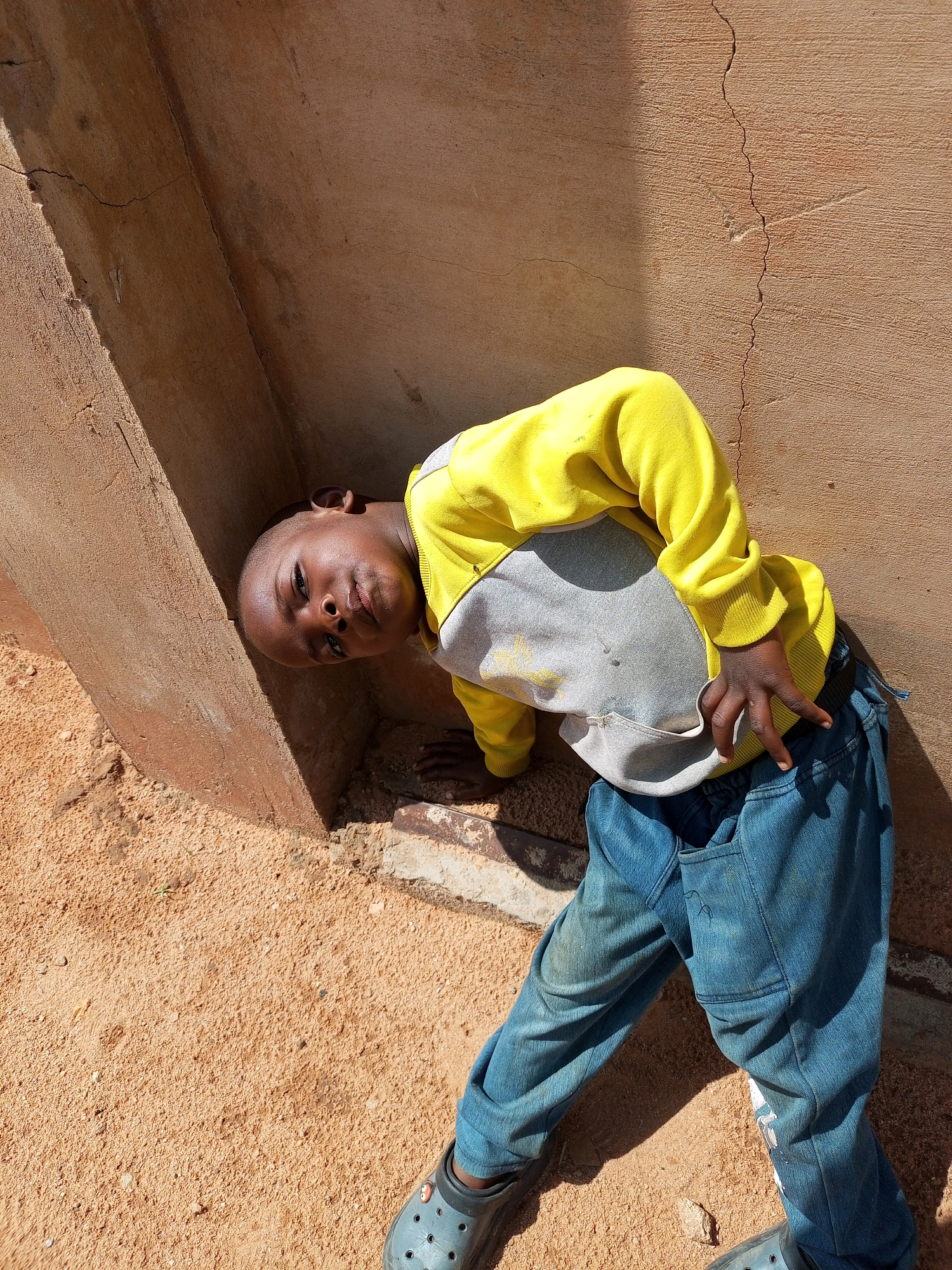 A young boy, appearing to be between 6 and 8 years old, is positioned in a corner formed by a rough, textured, terracotta-colored wall and a more heavily textured, tan-colored concrete structure. The boy is leaning into the corner, with his head tilted back and his body bent. He is wearing a yellow long-sleeved shirt with a grey section across the chest and blue denim jeans. His right hand is resting on the ground in front of him, while his left arm is bent and his hand is raised, with his index and middle fingers extended. His feet are clad in black slip-on shoes with visible strap detailing. The ground is covered in a coarse, sandy material, with some small stones and debris. The wall shows signs of weathering and has several cracks. The lighting suggests a sunny day, with distinct shadows cast by the boy and the structures. The location is described as being in Tempure, Nigeria.