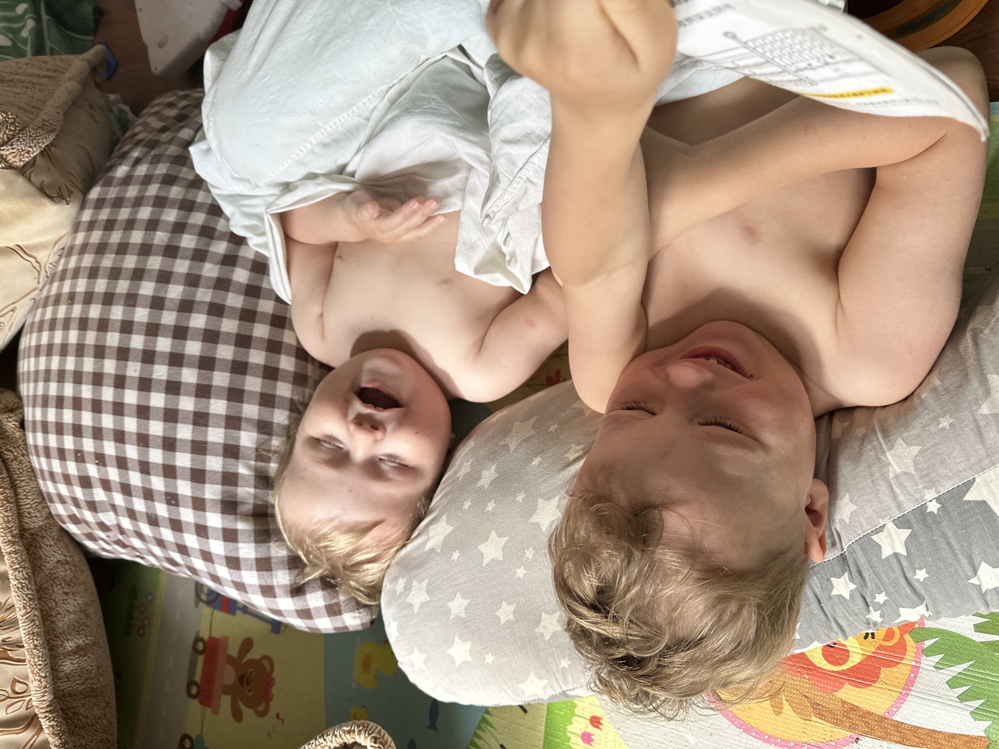 Two blond-haired children are lying on patterned pillows on a colorful play mat. The child on the left is partly covered by a white cloth and is looking upwards with their mouth open. The child on the right is holding a white instruction manual above them and has their eyes closed. The scene is indoors. There is no information in the image to suggest this location is Nizhny Novgorod, Russia.