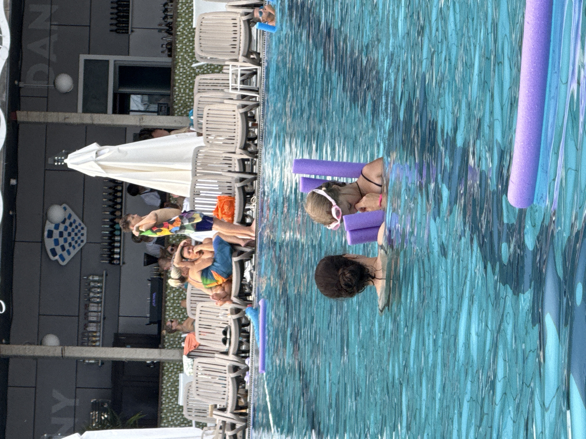 In the city of Yalta, Ukraine, a clear, turquoise swimming pool occupies the foreground. Two women are partially submerged in the water. The woman on the left has dark hair pulled back in a bun and is facing away from the camera. The woman on the right, a child wearing pink-rimmed goggles, is facing the camera and holding a purple pool noodle. Behind the pool, lounge chairs are arranged in rows, with several people relaxing on them. A bar area with bottles of alcohol is visible in the background, and a white umbrella provides shade. The overall scene depicts a leisurely day at a poolside establishment.