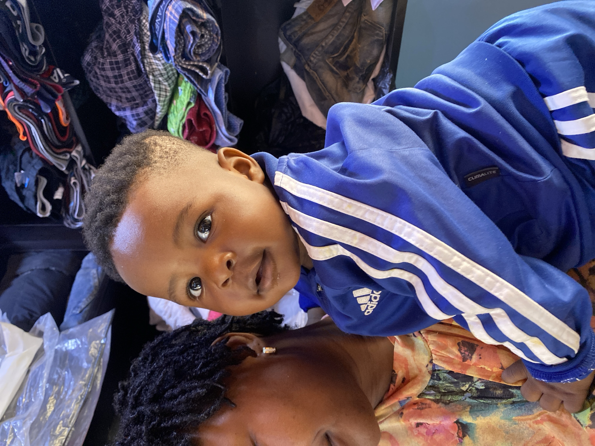 A close-up, angled shot depicts a young child in a blue Adidas tracksuit lying on top of an adult. The child has dark hair, brown skin, and is looking up at the camera. The adult has dark skin and dark hair with dreadlocks. The background consists of various folded clothes and jeans. The location is suggested to be Bukuru, Nigeria.