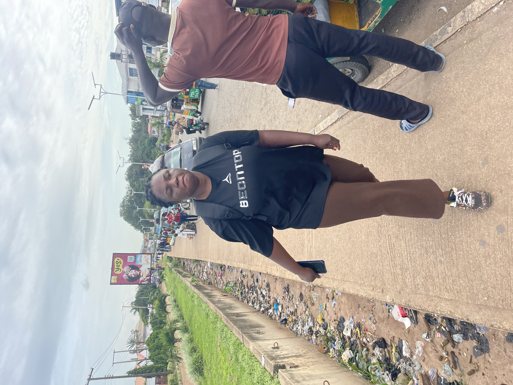 In Barakin Lerawi, Nigeria, a woman with dark hair pulled back walks on a concrete sidewalk. She is wearing a black t-shirt with white text, brown pants, and black and colorful athletic shoes. She carries a black backpack and holds a dark-colored smartphone in her right hand. To her right, a man with dark skin, wearing a brown t-shirt with white trim, dark pants, and light blue sandals, walks with his right arm behind his head. The sidewalk is bordered on the left by a concrete drainage ditch filled with litter. Beyond the ditch, there is a grassy area with palm trees. In the background, a billboard displays the word "yes!" and a QR code. Several vehicles, including a car, a motorcycle, and a three-wheeled auto-rickshaw, are visible on the road. The sky is overcast with scattered clouds.