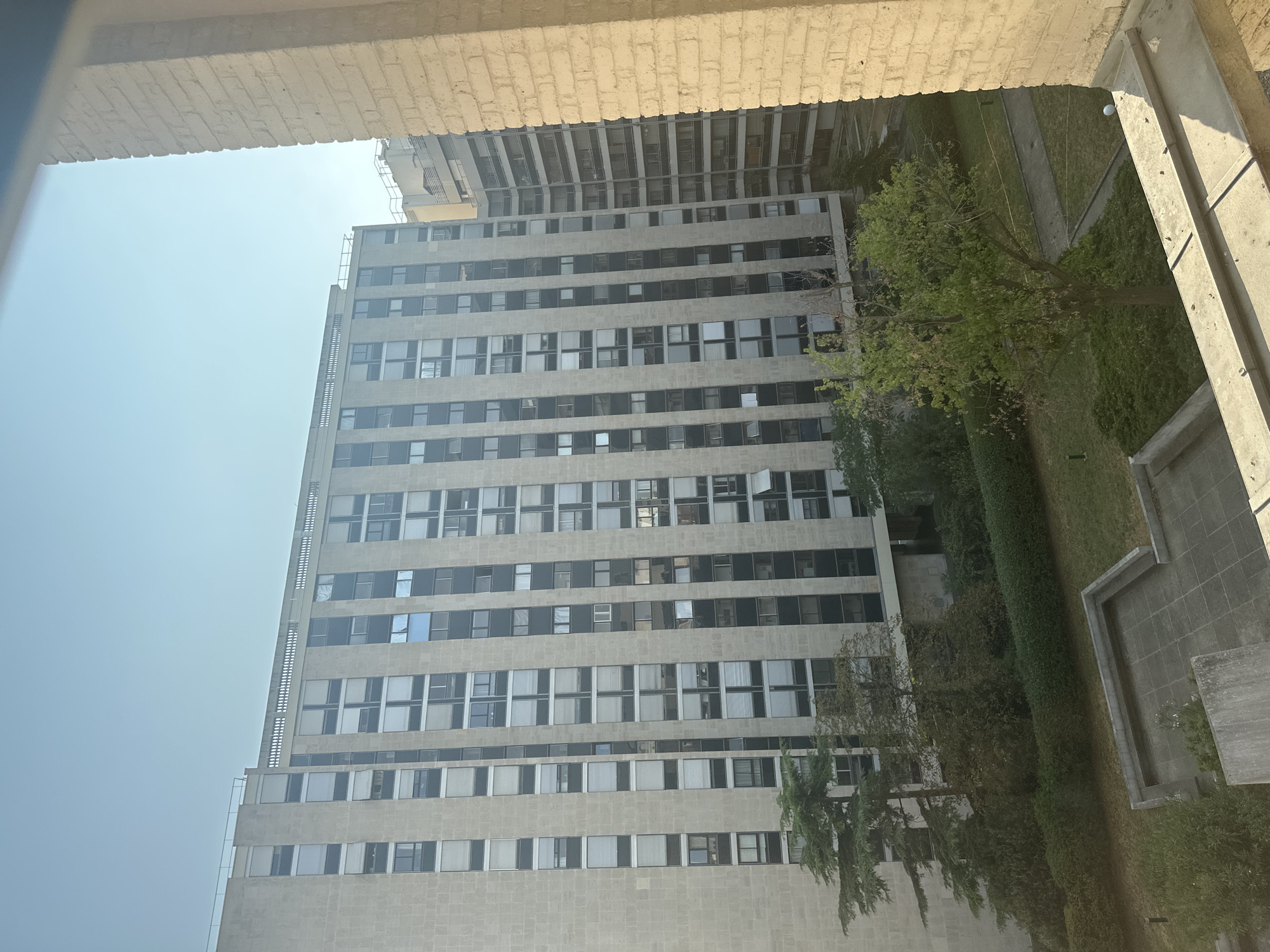 The image shows a view of a tall, modern apartment building in Paris, France, likely in a residential area. The building is constructed with a light-colored stone facade and features many windows, some of which are covered by blinds. The building appears to be of a brutalist architectural style, characterized by its large, imposing structure and geometric design. In front of the building, there is a green space with trees and hedges, as well as a paved area with concrete seating. The sky is clear and blue, indicating a sunny day. The perspective suggests the photo was taken from another building or a higher vantage point.