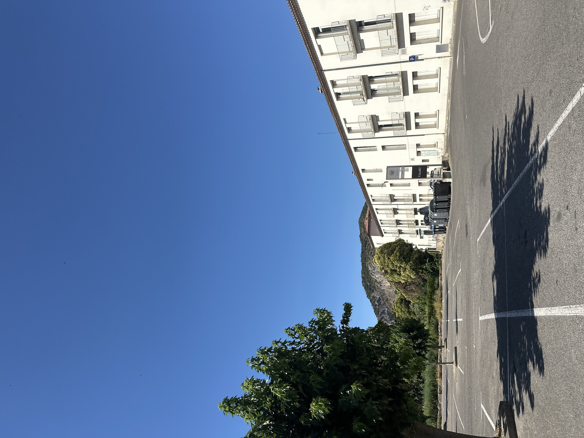 The image shows an empty parking lot in front of a white, multi-story building in Anduze, France. The building has several windows and balconies. To the left of the building, there is a large green tree. In the background, there are hills and more trees. The sky is clear and blue. There are trash bins lined up in front of the building.