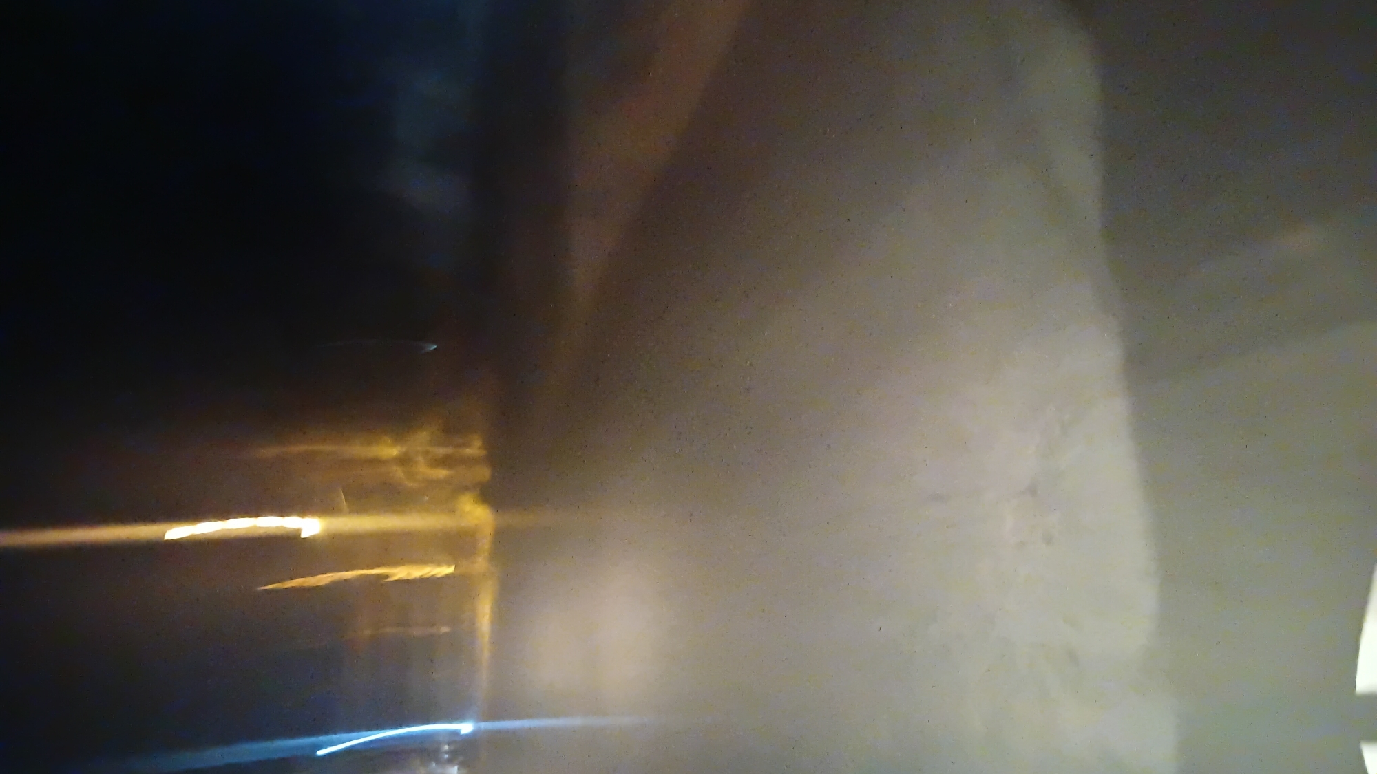The image depicts a blurred nighttime scene, likely captured from a moving vehicle. The foreground is dominated by a light-colored, textured surface, possibly a road or pathway, illuminated by a bright, diffused light source. In the background, streaks of light suggest artificial lighting from streetlamps or buildings, with distinct yellow and blue hues. The overall impression is one of motion and low light conditions. While the image is abstract and lacks specific identifiable landmarks, the presence of streetlights and the texture of the ground suggest an urban or semi-urban environment. It is not possible to definitively determine the specific location within Prabumulih, Indonesia, based on this image alone.