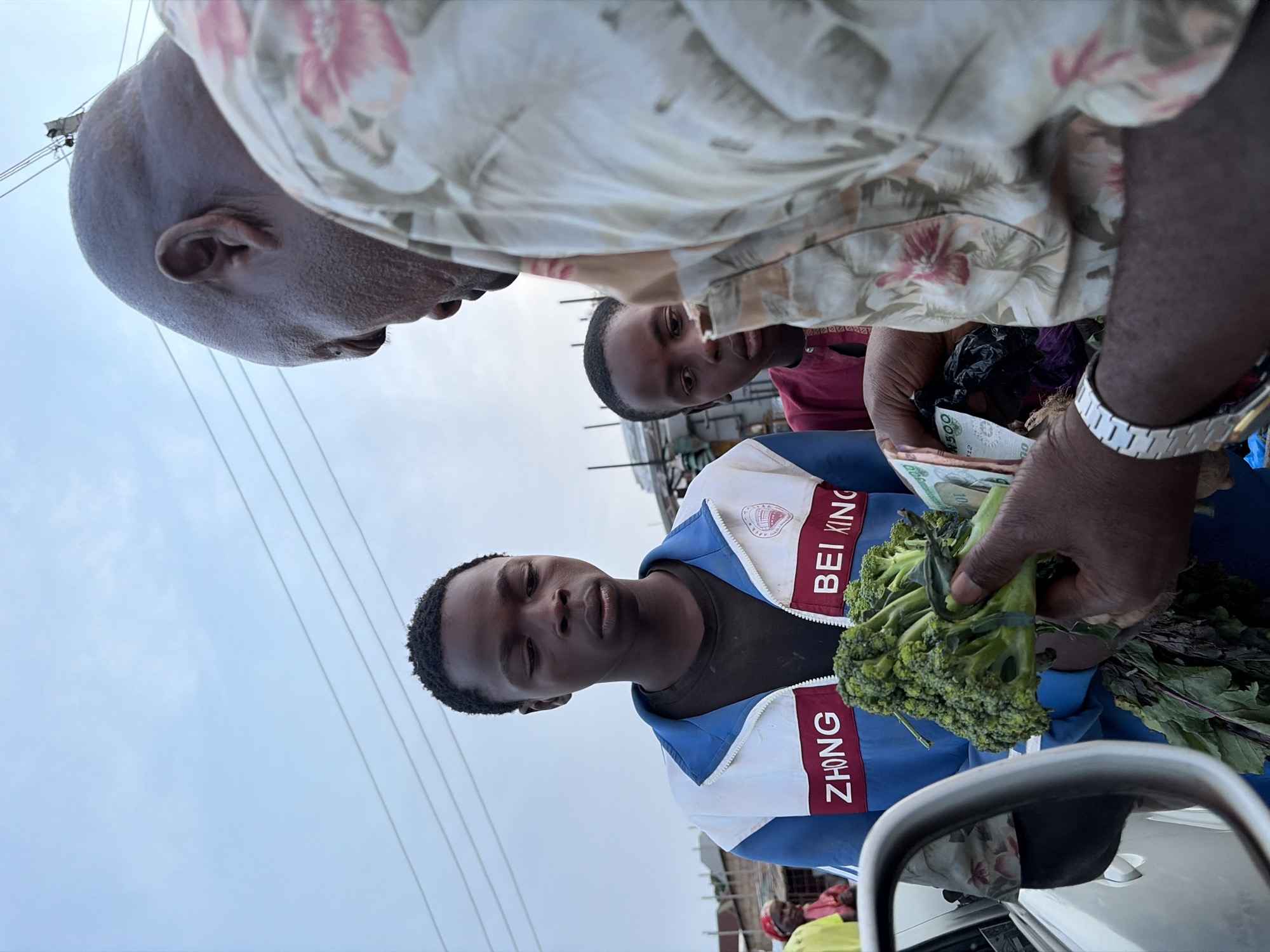 In Gura Top, Nigeria, an outdoor scene depicts a bald, dark-skinned man in a floral shirt, examining a bundle of broccoli held by a dark-skinned person in a blue and white jacket labeled "BEI KING" and "ZHONG". Currency is visible in the man's hand. A third person looks on from behind the man, adding to the interaction occurring. The edge of a vehicle's side mirror is visible in the foreground. Overhead wires are present against a pale sky.