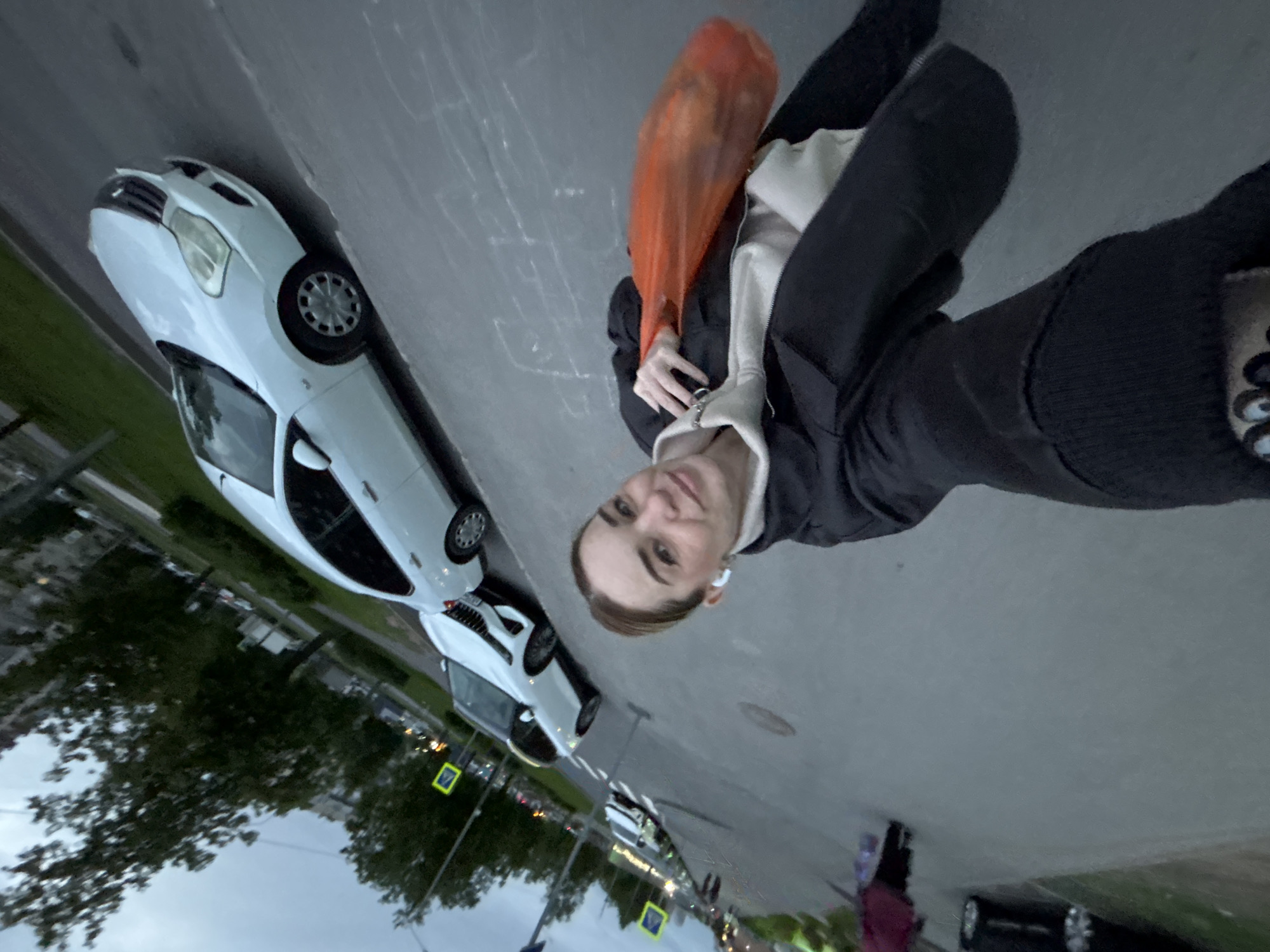 A woman poses for a selfie outdoors with a white car behind her, alongside a city street in the city of Unknown, Unknown. She wears a dark jacket, a white top, and black socks with a white design on the toes. She also holds an orange plastic bag. The location is urban, featuring an asphalt street lined with parked cars, trees, and street signs. The sky is overcast, suggesting a cloudy day.