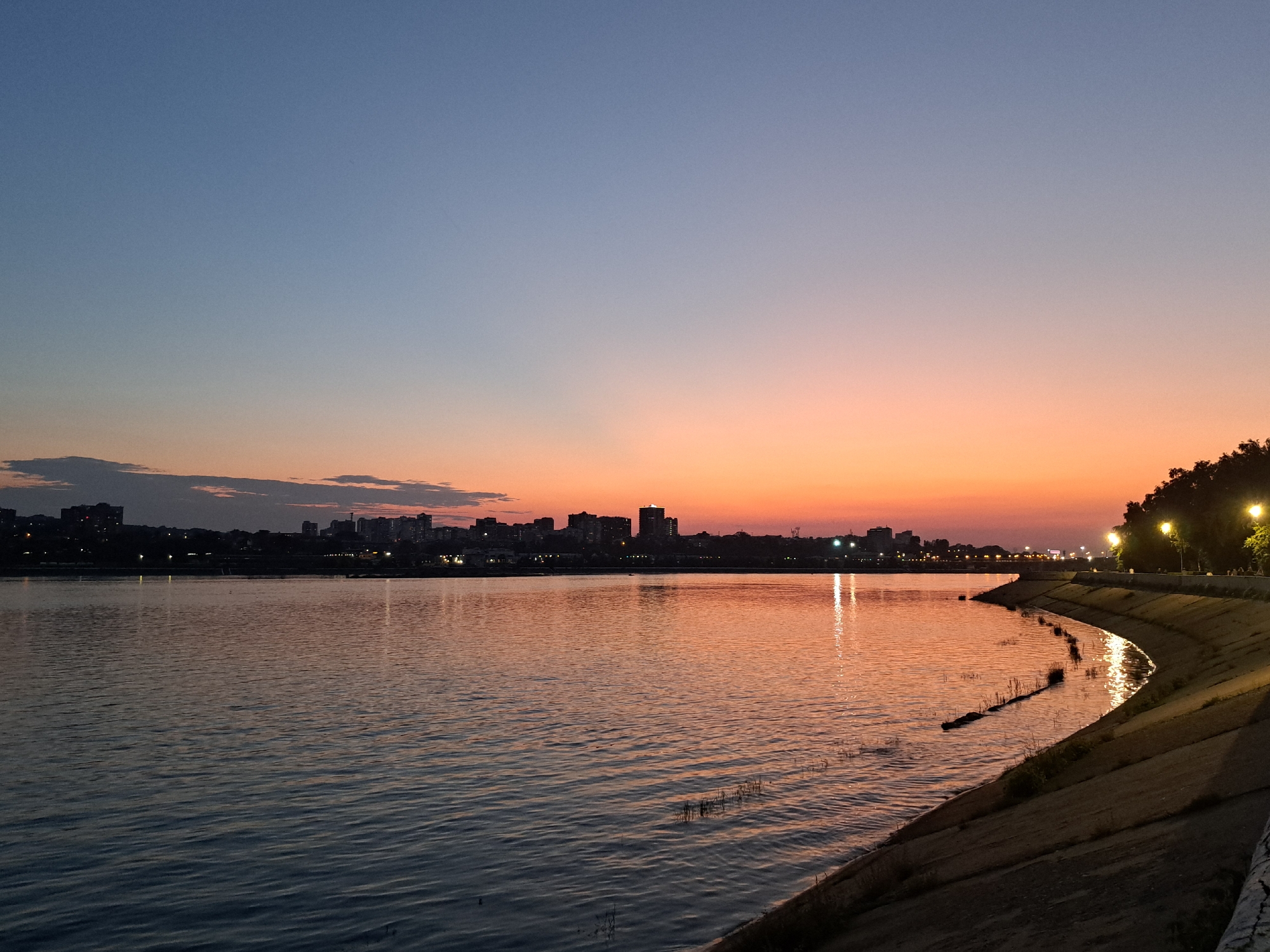 The image depicts a serene sunset over the Angara River in Irkutsk, Russia. The sky transitions from a clear, pale blue at the top to a warm gradient of orange and pink near the horizon, with a few wispy clouds visible. Across the river, the silhouette of the Irkutsk city skyline is visible, with buildings of varying heights illuminated by the fading light. Numerous streetlights and building lights create a soft glow along the waterfront.
The foreground features the calm surface of the Angara River, reflecting the colors of the sunset and the city lights. On the right side of the image, a concrete embankment curves along the river's edge, with a path and streetlights lining the top. The embankment is composed of layered concrete sections, showing signs of wear and tear. Sparse vegetation is visible growing between the concrete slabs and at the water's edge. The scene is tranquil, with no people or specific actions clearly discernible. The overall impression is one of a peaceful urban landscape at dusk.