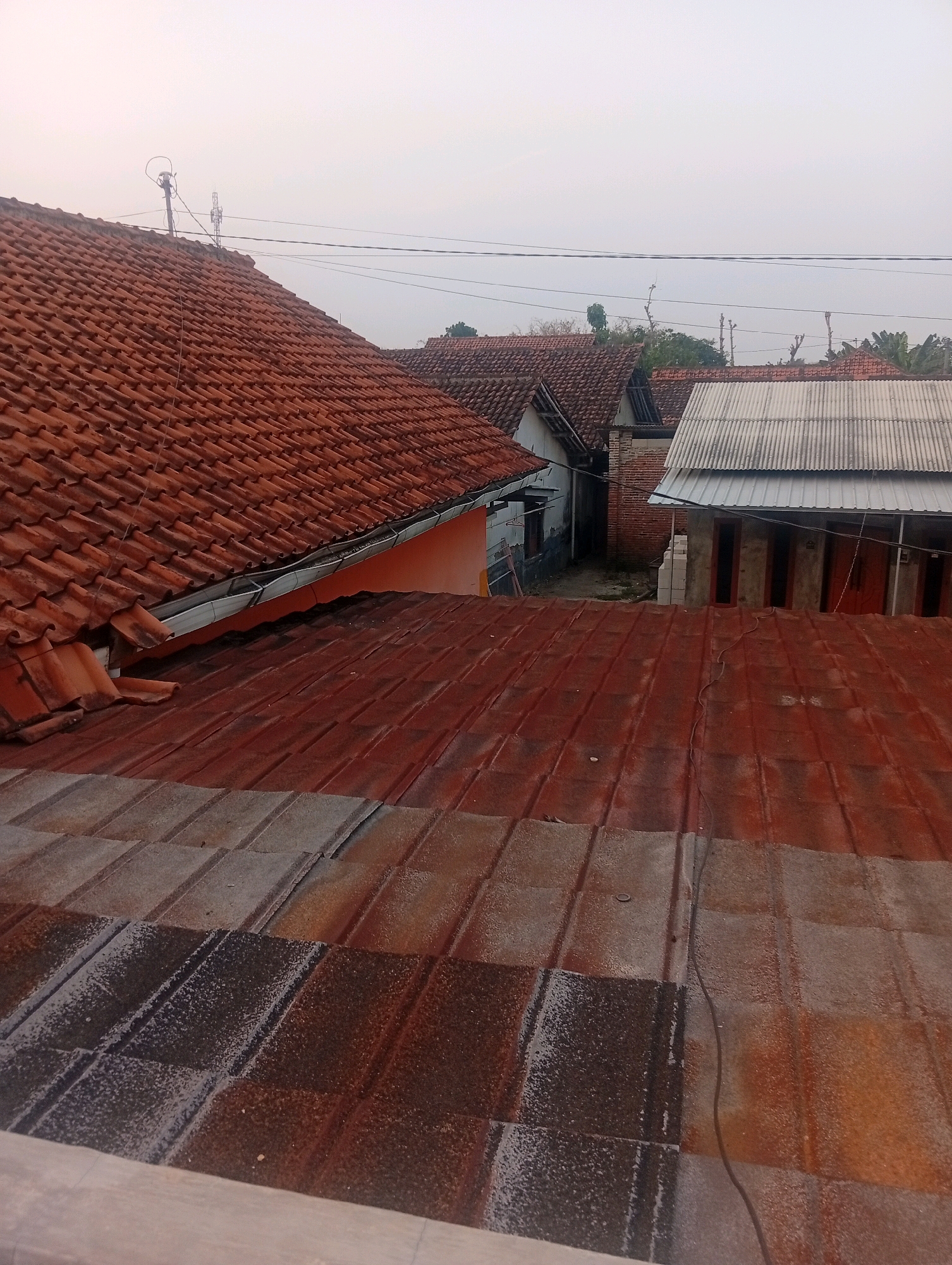 The image captures an overhead view of rooftops in the Kebandingan, Indonesia area. The foreground is dominated by a rusty, red metal roof with distinct parallel ridges. Further back, the scene transitions to clay tile roofs, primarily in a reddish-brown hue. The roofs show varying states of wear, with some appearing older and more weathered than others. A white corrugated metal roof is visible on a structure to the right, contrasting with the dominant red tones. Behind the rooftops, a few buildings with brick and plaster walls are partially visible, along with glimpses of trees and foliage. The sky is overcast with a pale, hazy light, suggesting either early morning or late afternoon. Power lines are strung across the upper portion of the image, connecting to poles visible on the rooftops. The overall impression is of a residential area characterized by traditional roofing materials and modest structures.
