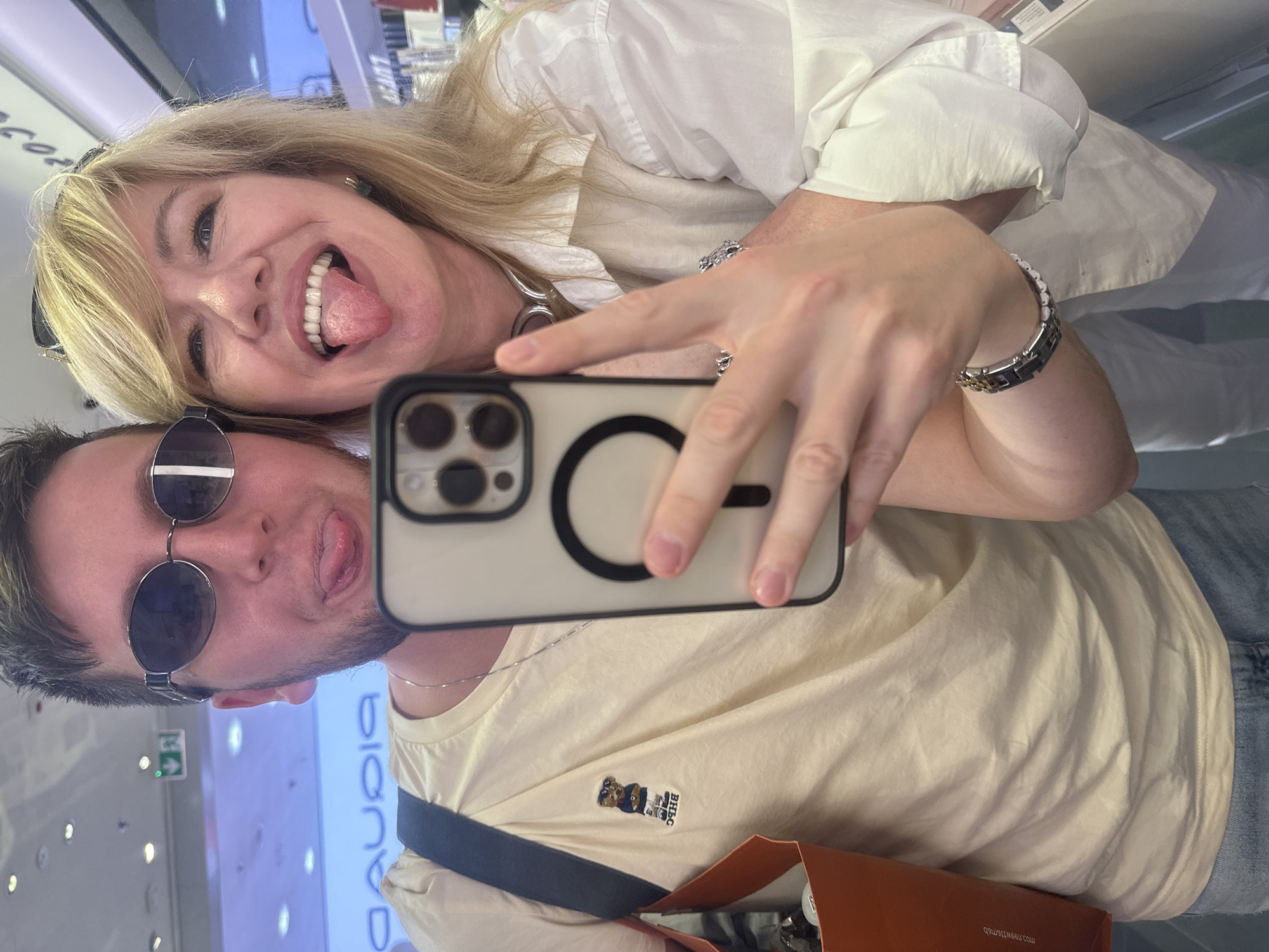 The media file is a selfie taken by two people, a woman and a man, with a phone. They are both sticking their tongues out playfully. The woman has blonde hair and is wearing a white shirt. The man has short, dark hair and is wearing sunglasses and a cream-colored t-shirt with a small bear graphic. The background appears to be an indoor retail environment with shelves and displays, possibly a store. There is text visible on a blue sign in the background which reads "BLANCA", and on an orange bag in the foreground which reads "mosnow.newsmob". The image conveys a lighthearted and fun emotion. The time of day and weather are not discernible. The context is Moscow, Russia.