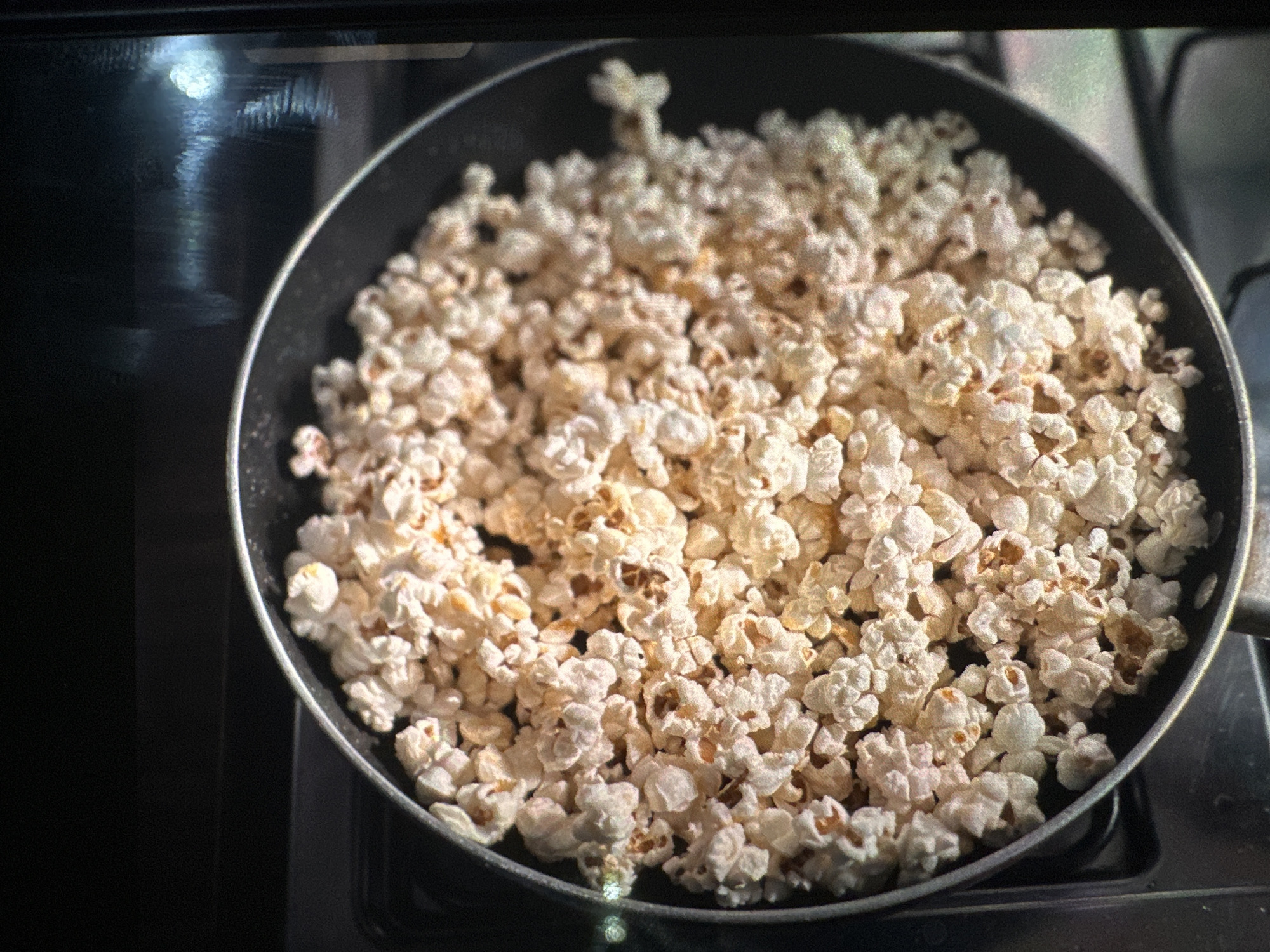 An overhead close-up shot shows a black frying pan filled with freshly popped popcorn. The popcorn is light in color with some darker, toasted kernels interspersed throughout. The pan appears to be sitting on a stovetop, indicated by a dark surface with some faint reflections. The background is mostly dark and out of focus. This image does not depict any specific location in the city of Rido, Nigeria.